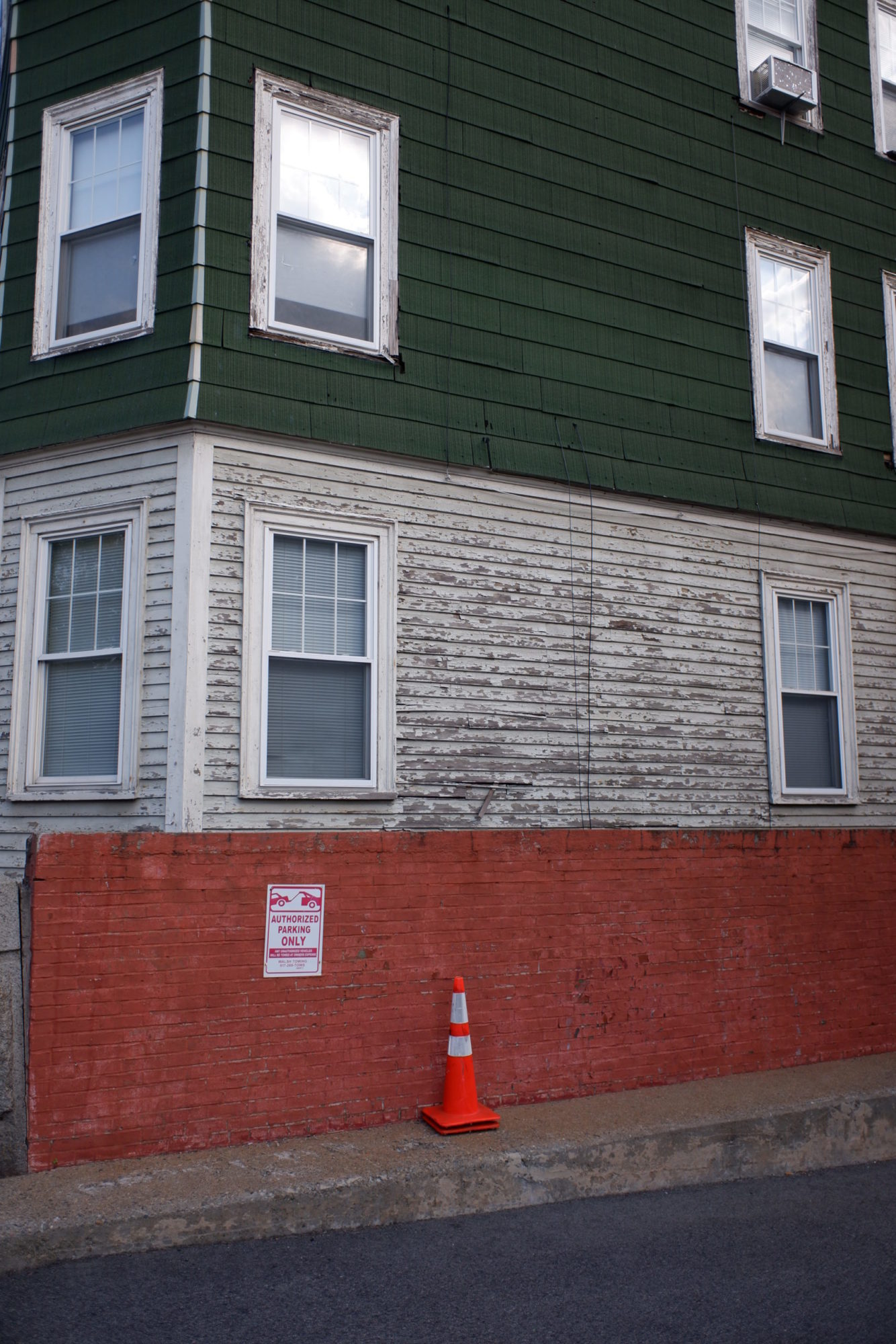 A closer shot of the side of a building. The bottom floor has a red brick wall against it. The second floor is older white wood. Stories above are dark green. Multiple windows are visible, one of which has an air conditioning unit in it. There is an orange cone on the sidewalk.