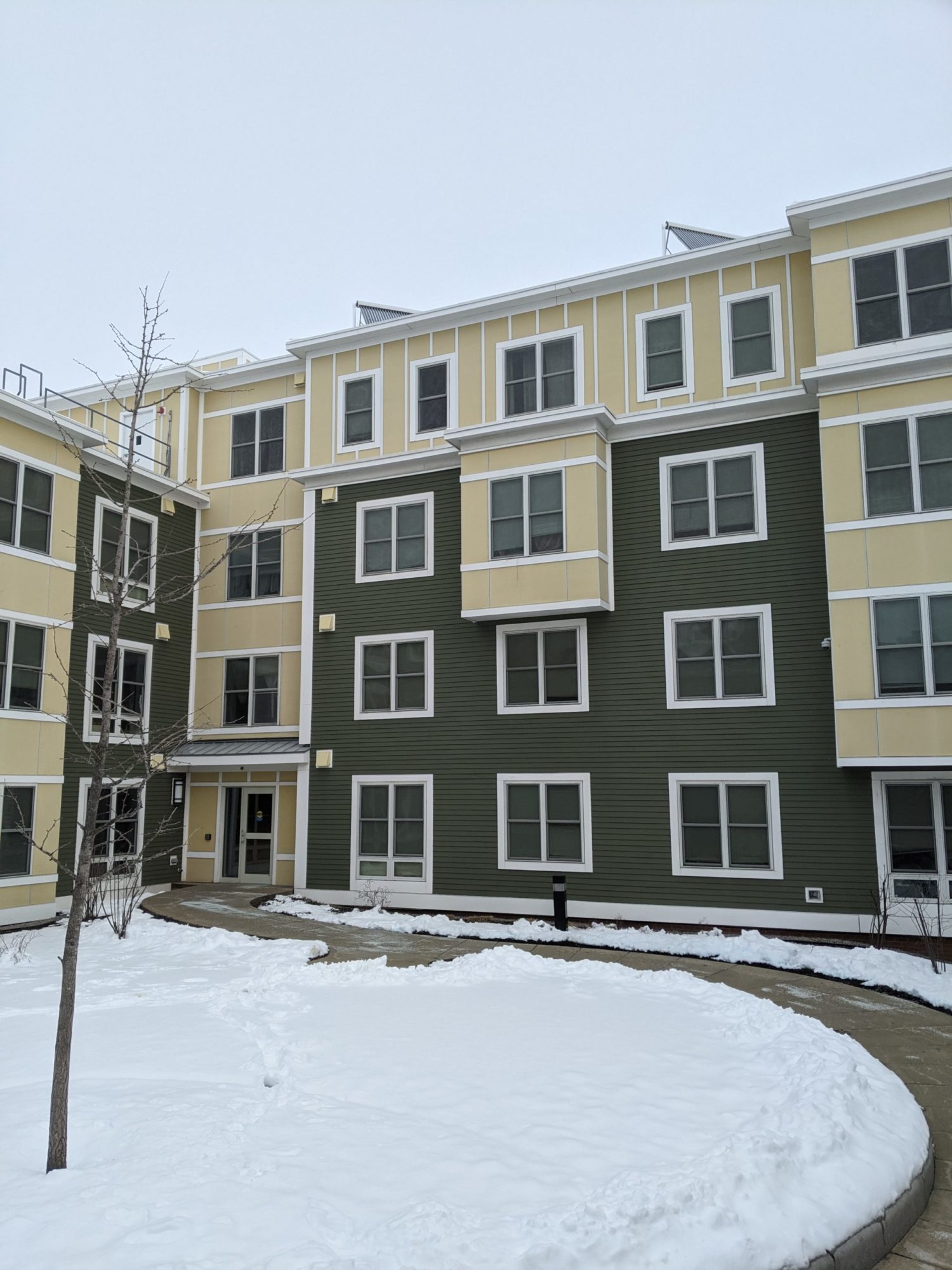 An angled view of an apartment building. A curved side walk cuts through the courtyard covered in snow. The sidewalk has been shoveled of snow. The building is a muted yellow and an olive green. It is unclear how many apartments are in view. There are two entrance doors and many windows in view.