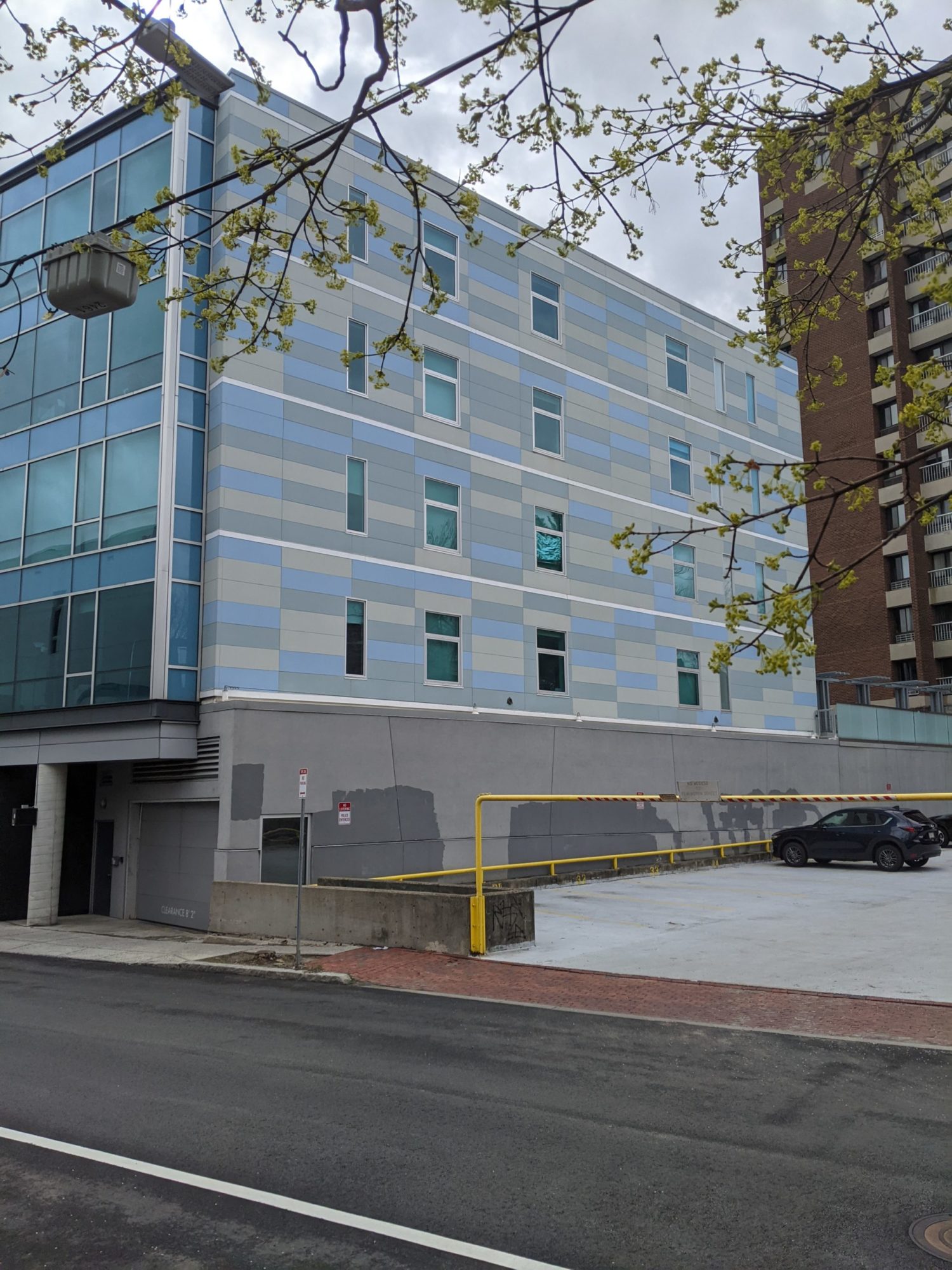 An angled shot of a multi story apartment building and the top floor of a parking deck. The building has four floors in view. The front is glass, the side is cinderblocks varying between light blue, light grey, and light taupe. Another apartment building is in view.
