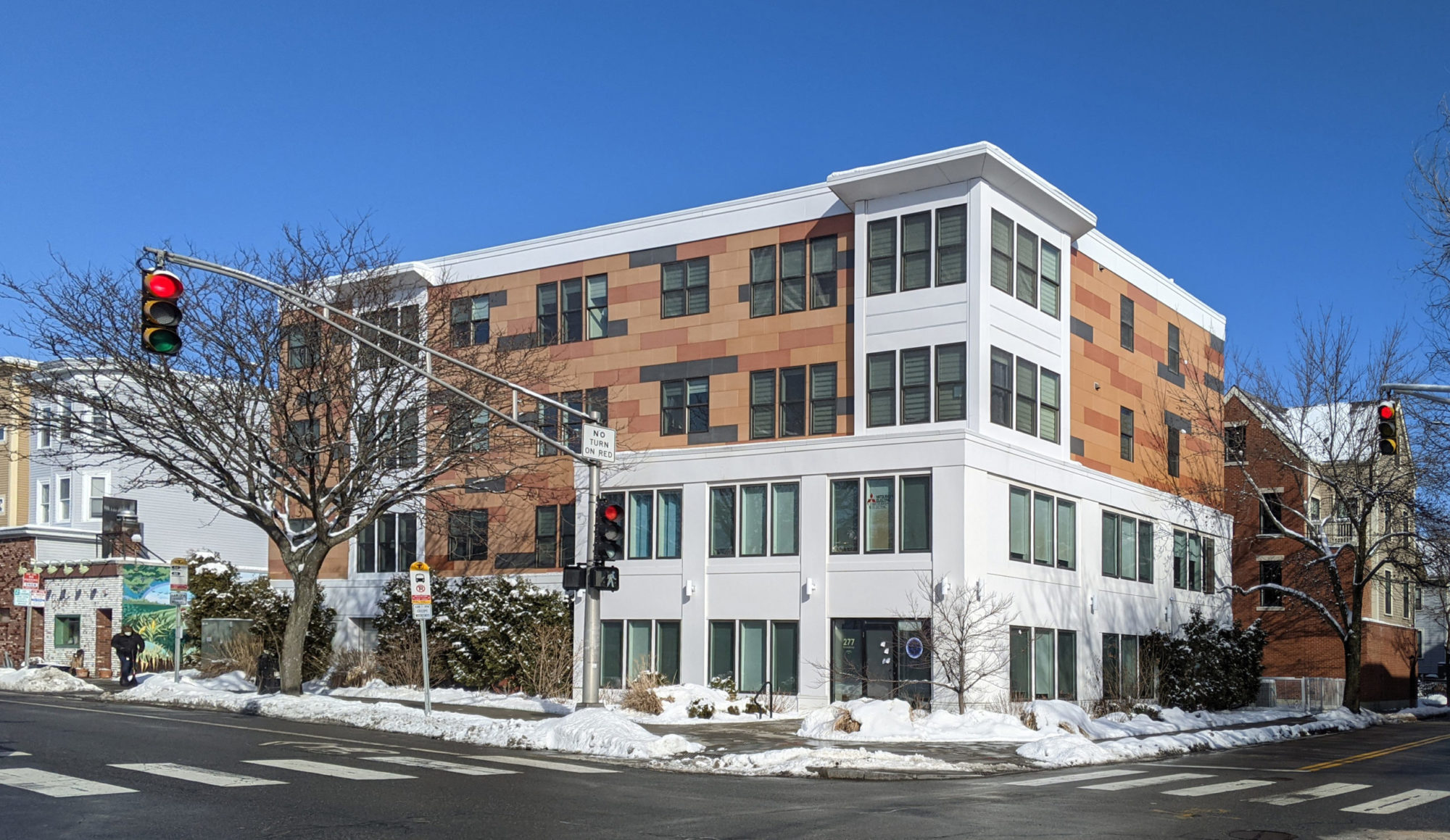 An angled, across the street shot of an apartment building. The building is white concrete and varying cinderblocks of different warm browns. There are many windows and one entrance door in view. There are multiple street lights in view. The road has been plowed for snow.