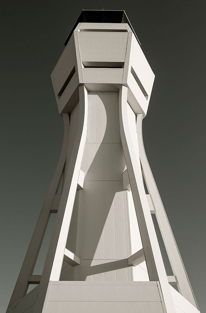 A white, geometric structure is photographed from a low angle. It extends vertically into a grey sky.