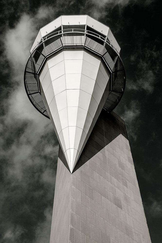 A white structure comes to a sharp point. It appears attached on one side to a slim building. In the background, the sky appears cloudy.