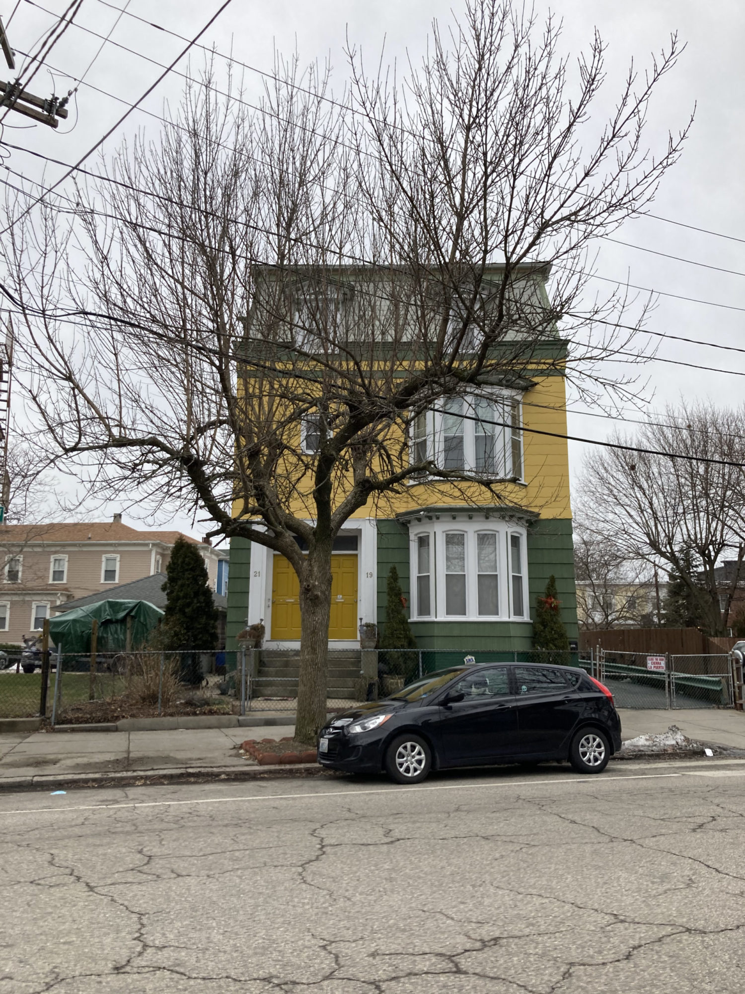 A straight on shot of a multi story house painted yellow and dark green. The front door is yellow and on the left side of the front of the house, above a few stairs. There is a tree directly in front of the house. There are two bay windows on different stories on the right side of the house.