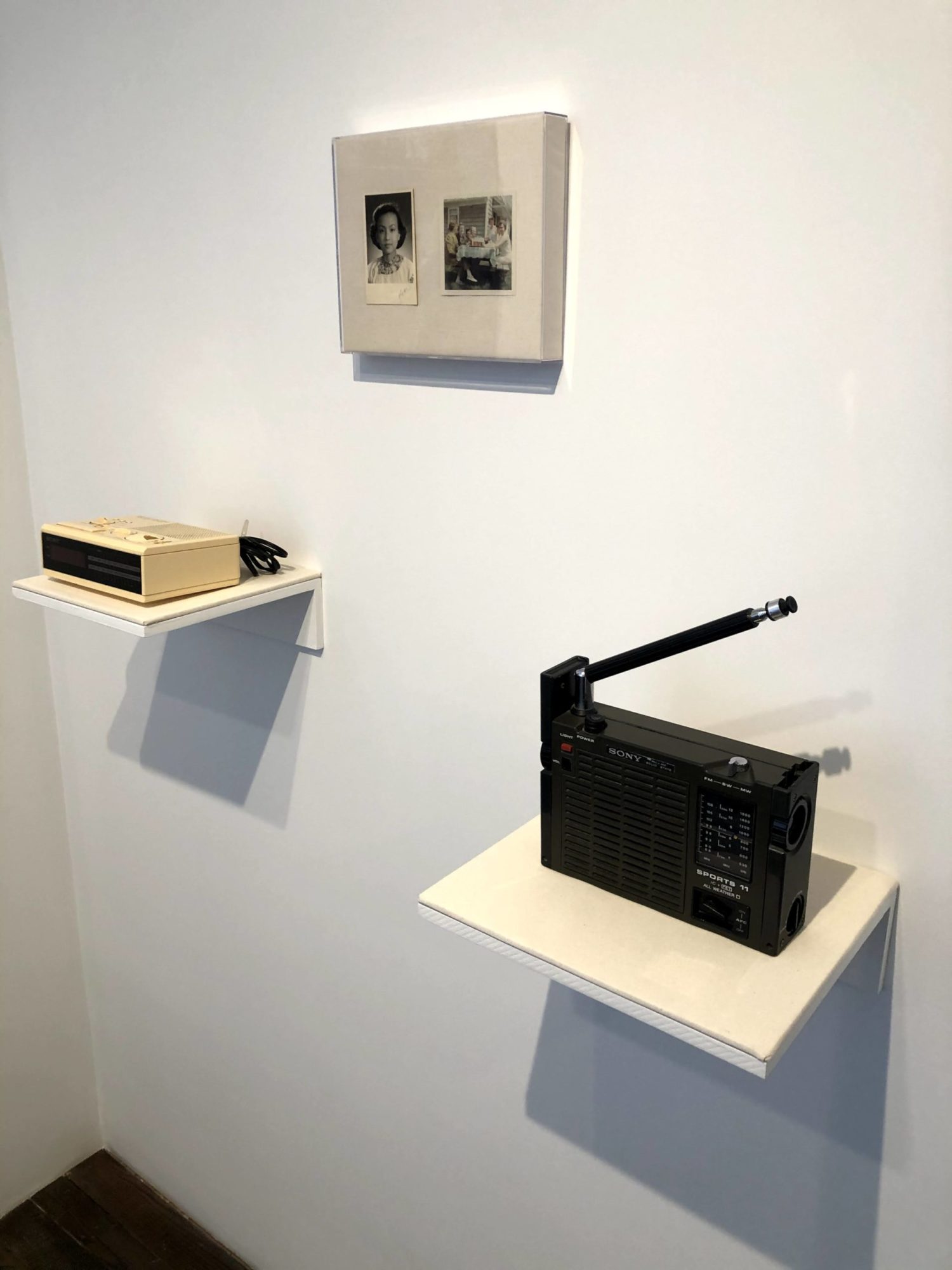 An angled installation shot. There are two square, floating shelves at staggered heights and two black and white images mounted together above. The floating shelves have antique radios on them; the higher shelf on the left featuring a small tan radio and the lower shelf on the right featuring a larger black radio with an antenna. The photo on the left above is a portrait and the photo on the right above is a group photo.