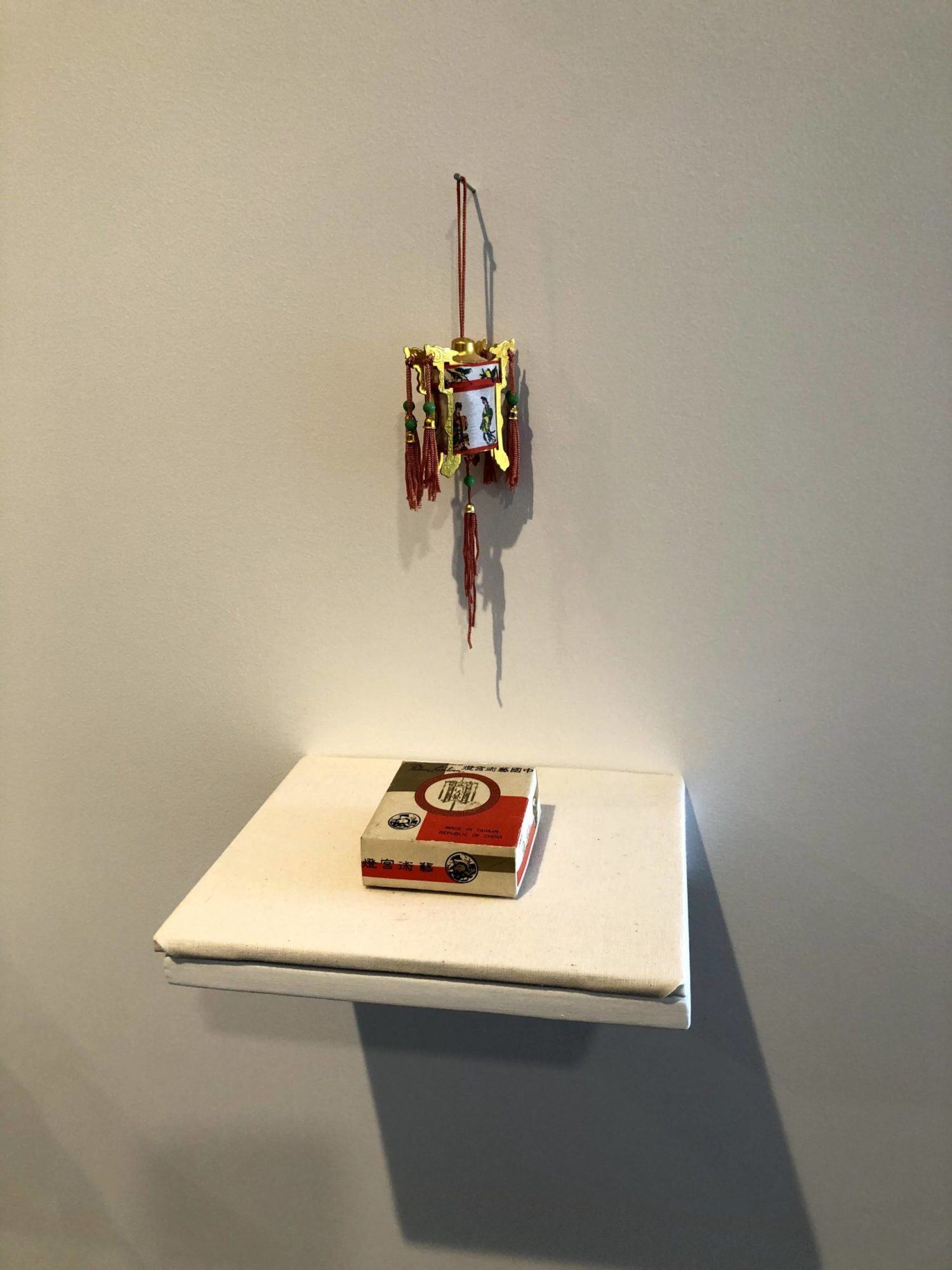 An installation view. There is a square floating self with a small, square, white, green, and red geometrically patterned box on it. Hanging on the wall above is small mixed media sculpture with red. green, and yellow tassels and beads.