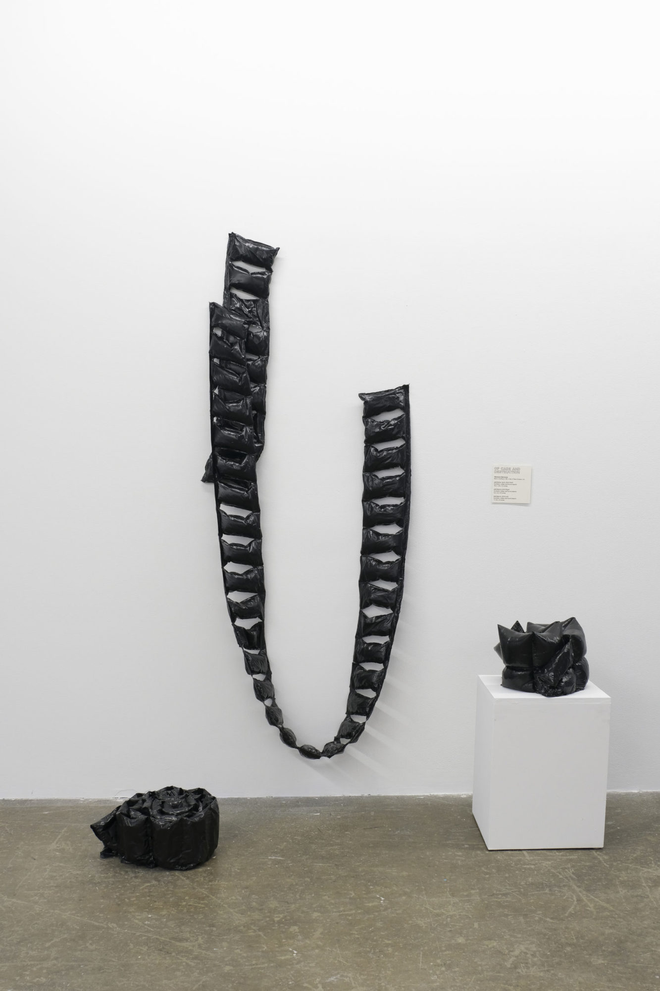 Installation view shows three synthetic rubber and found objects. One piece hangs from, one sits atop a pedestal, and a third sits on the ground.
