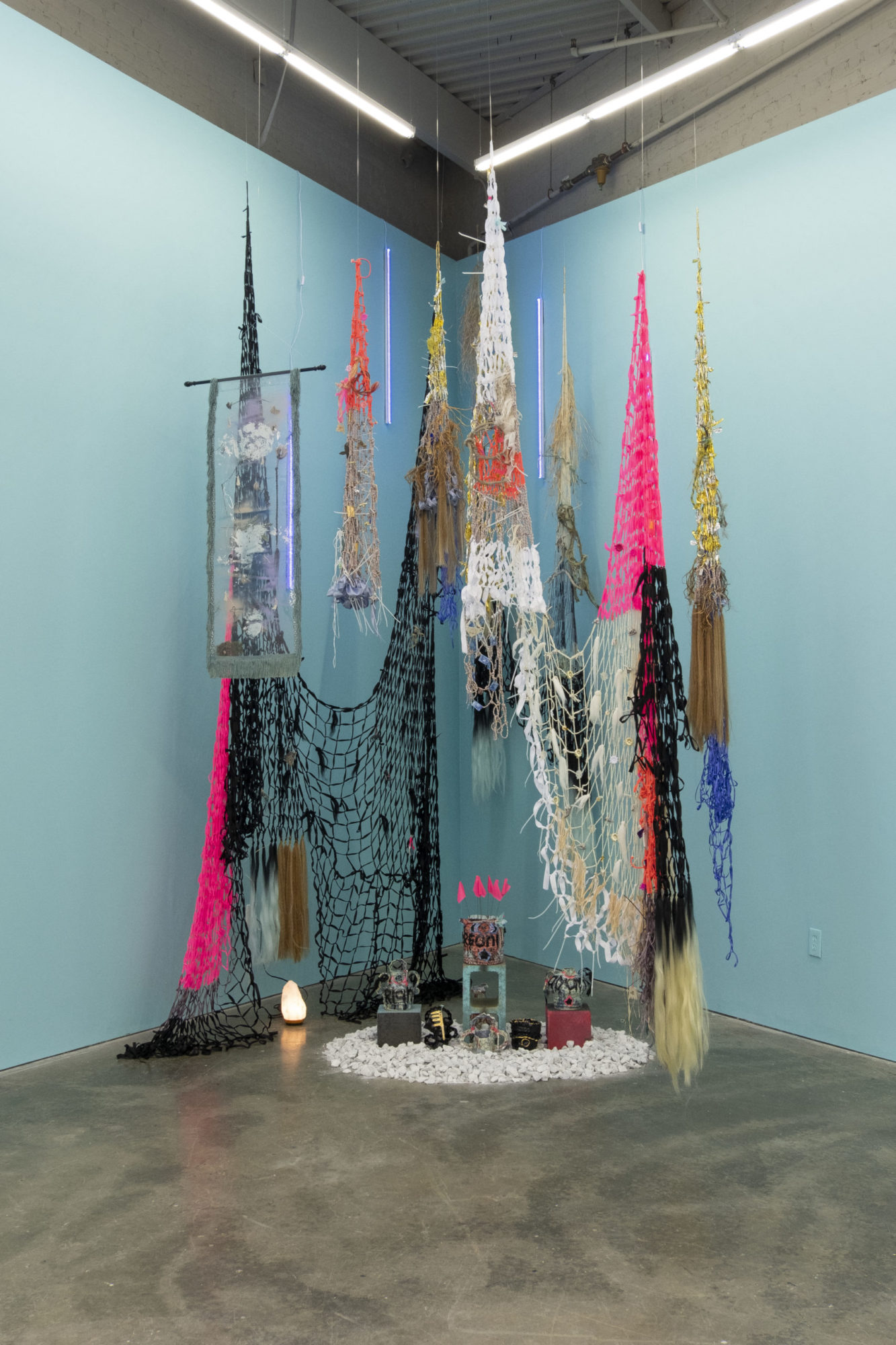 A mixed media sculpture, composed primarily of colorful textiles, is positioned in the corner of the gallery space.