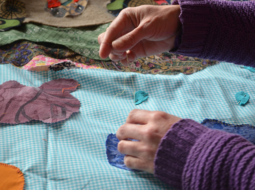 A close up photograph shows someone sewing a piece of fabric onto the textile, using yellow thread.