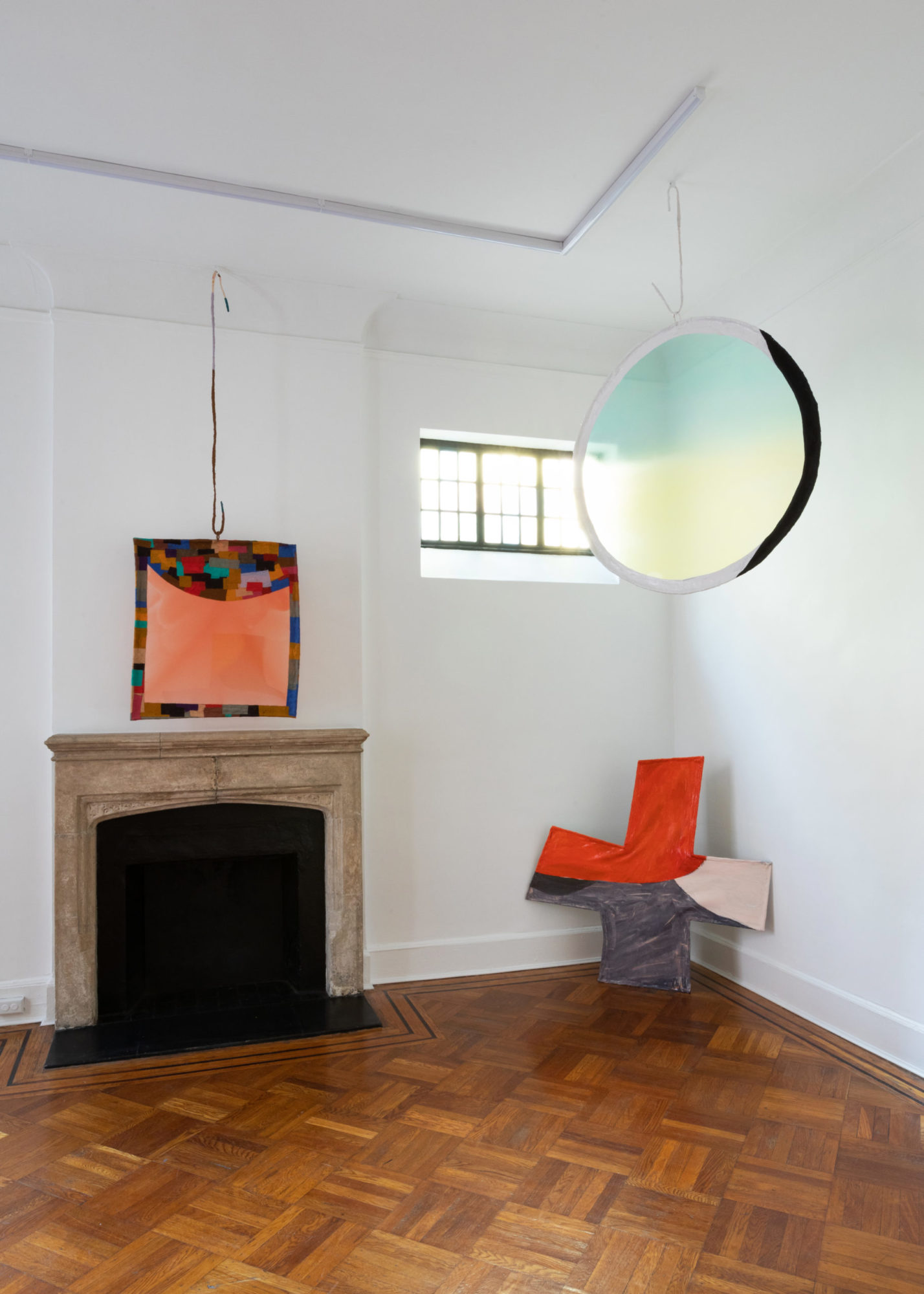 An installation view of Fabienne Lasserre's exhibition at TURN GALLERY in New York. Two pieces are hanging from the ceiling in a brightly lit room and one is propped against the wall.