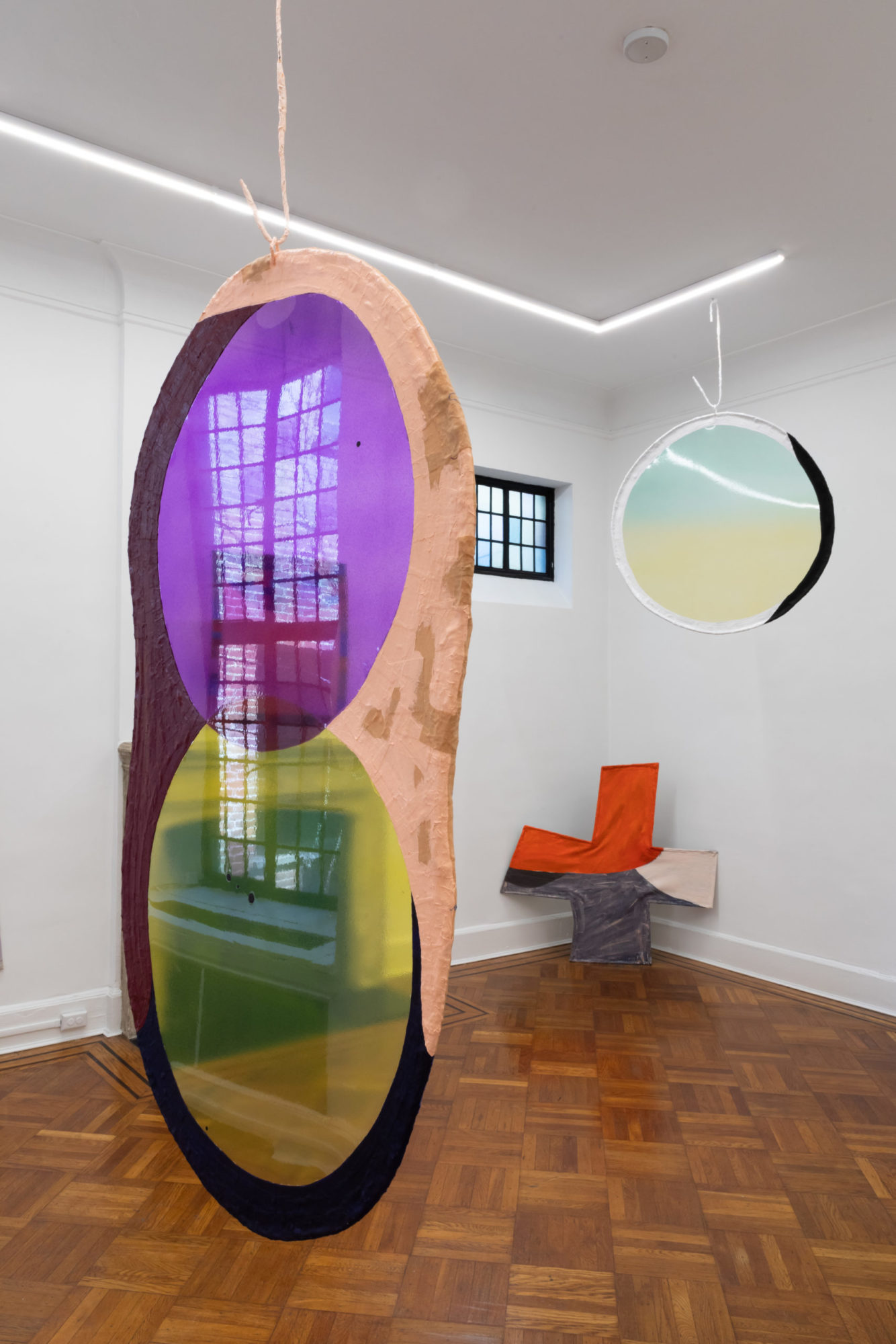 An installation view of Fabienne Lasserre's exhibition at TURN GALLERY in New York. Two pieces are hanging from the ceiling and a piece is propped against the wall. The circular pieces are vibrantly colored.