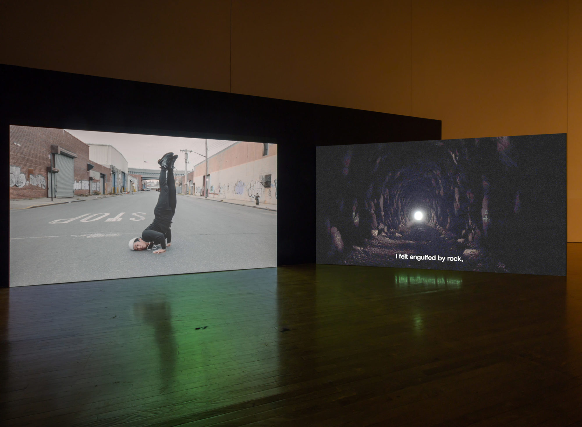 An installation view of Kenneth Tam's Silent Spikes at the Queen's Museum. Two screens are in view. The screen on the left features a man in the middle of an empty road doing a hand stand with the side of his face on te ground. he is wearing all black with a light beanie. The screen on the left features a shot through a cave with a light at the end. The text at the bottom reads 