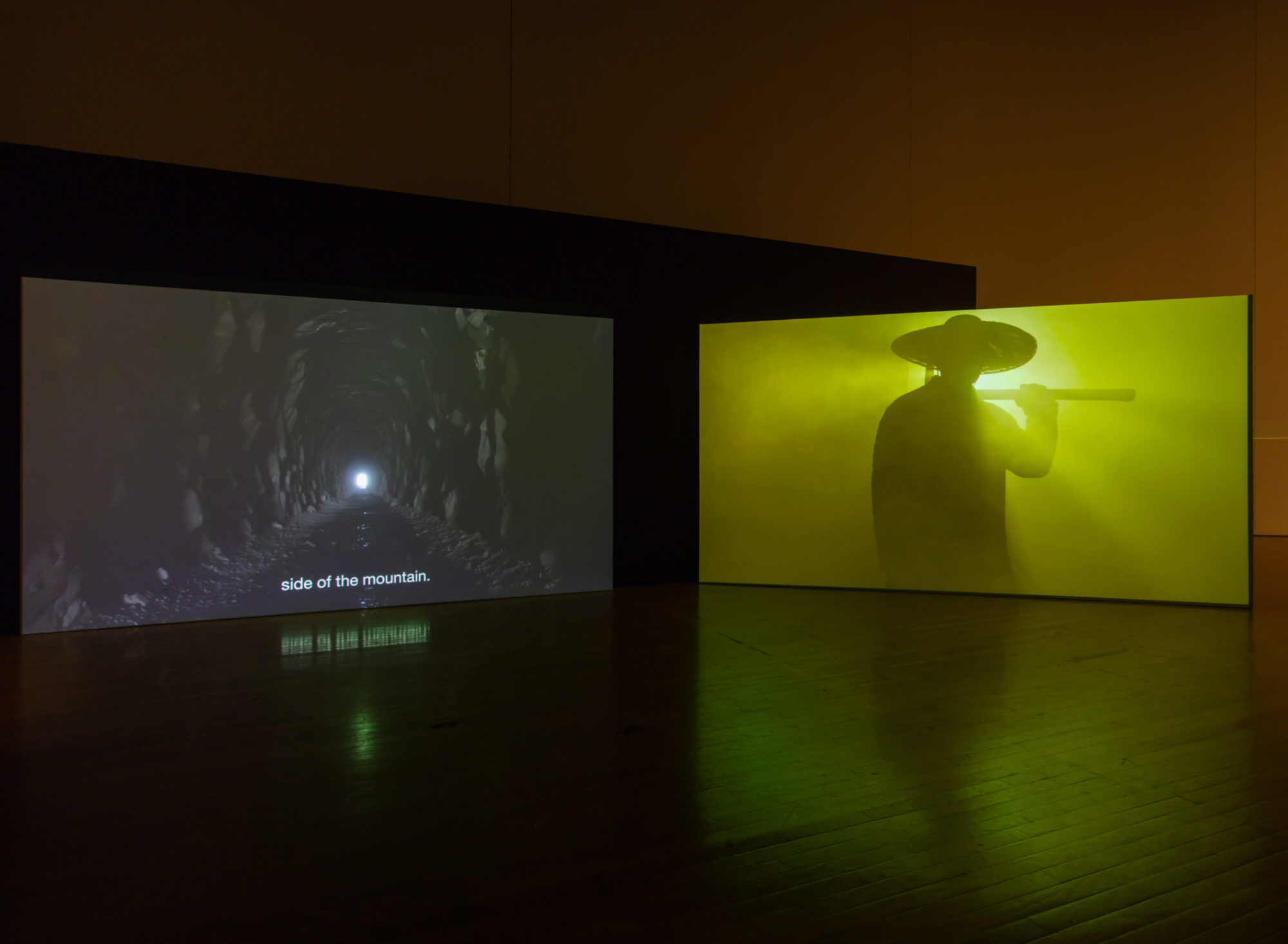 An installation view of Kenneth Tam's Silent Spikes at the Queen's Museum. Two screens are in view. The screen on the left is a view through a cave with a light at the end. The text at the bottom reads 