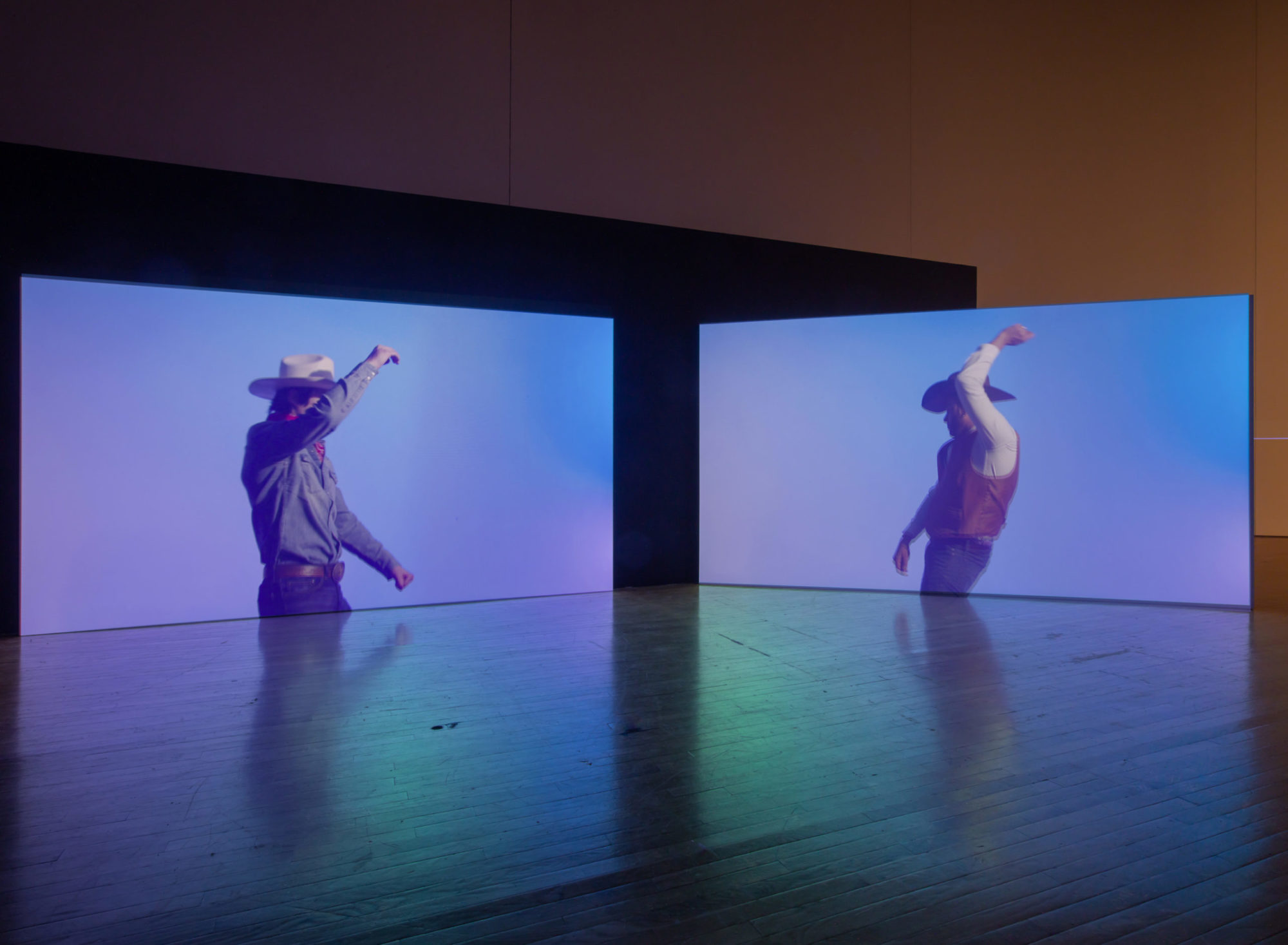 An installation view of Kenneth Tam's Silent Spikes at the Queen's Museum. Two screens are in view, both featuring cowboys dancing in front of a blurry blue and lavender background. Neither of their faces are in view. The lights in the gallery space are dimmed.