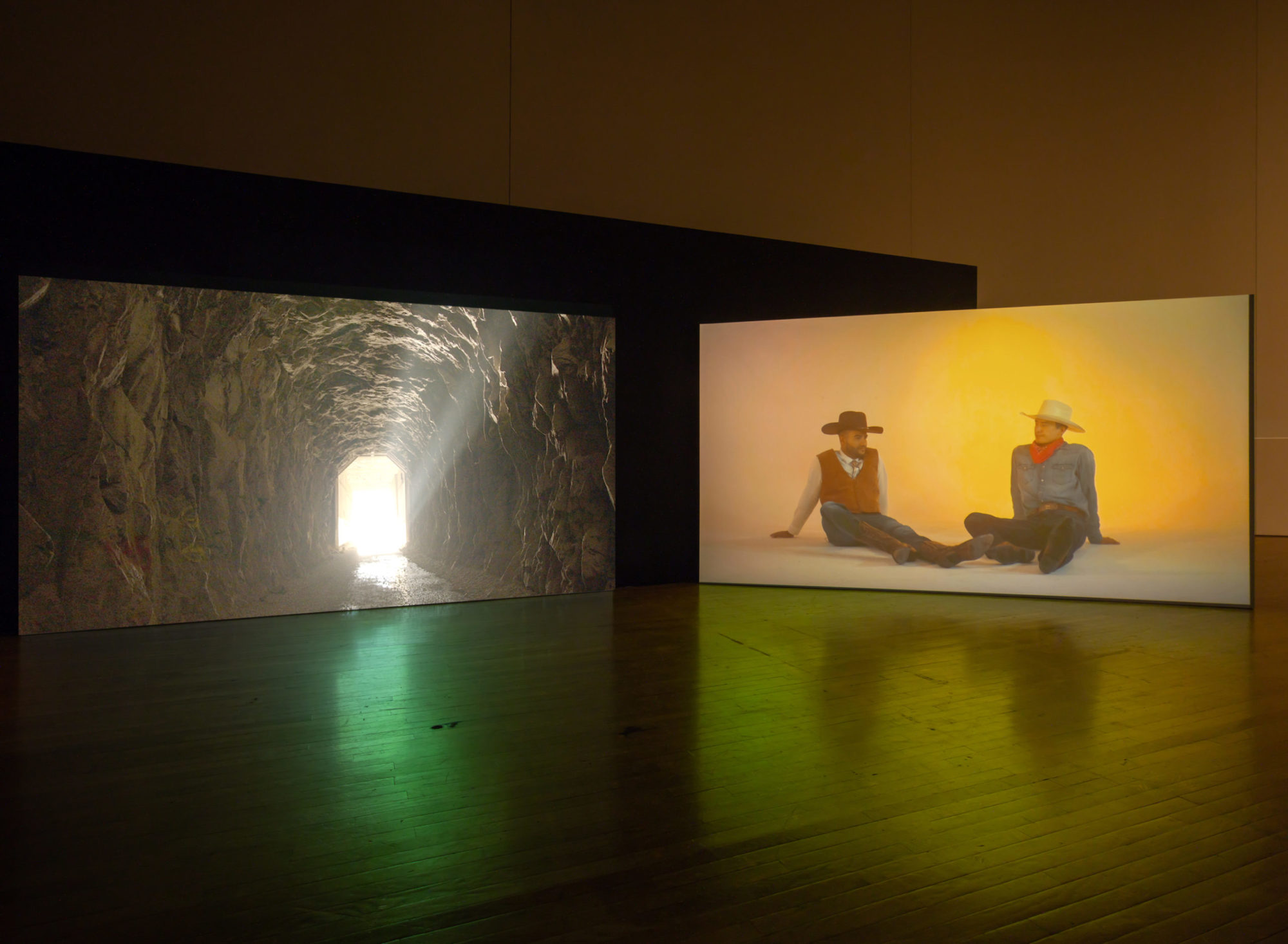 An installation view of Kenneth Tam's Silent Spikes at the Queen's Museum. Two screens are in view. The screen on the left is a view of the edge of a cave, the exit in view and light pouring in. The image on the right is two men sitting on the ground. The background is a warm orange yellow. The man on the left wears boots, jeans, a white shirt, a brown vest, and a dark hat. The man on the right wears boots, dark jeans, a chambray shirt, a red bandana around his neck, and a light hat.