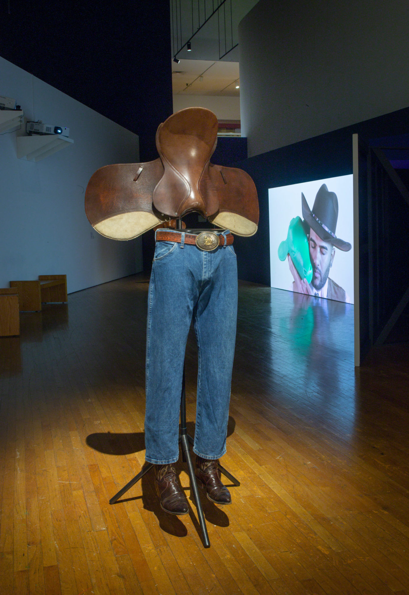 An installation view of Kenneth Tam's Silent Spikes at the Queen's Museum. The image features a sculpture of the legs of a mannequin clad in denim, cowboy boots, and a belt and the upper half being comprised of a brown leather saddle. The lights in the gallery space are dimmed.