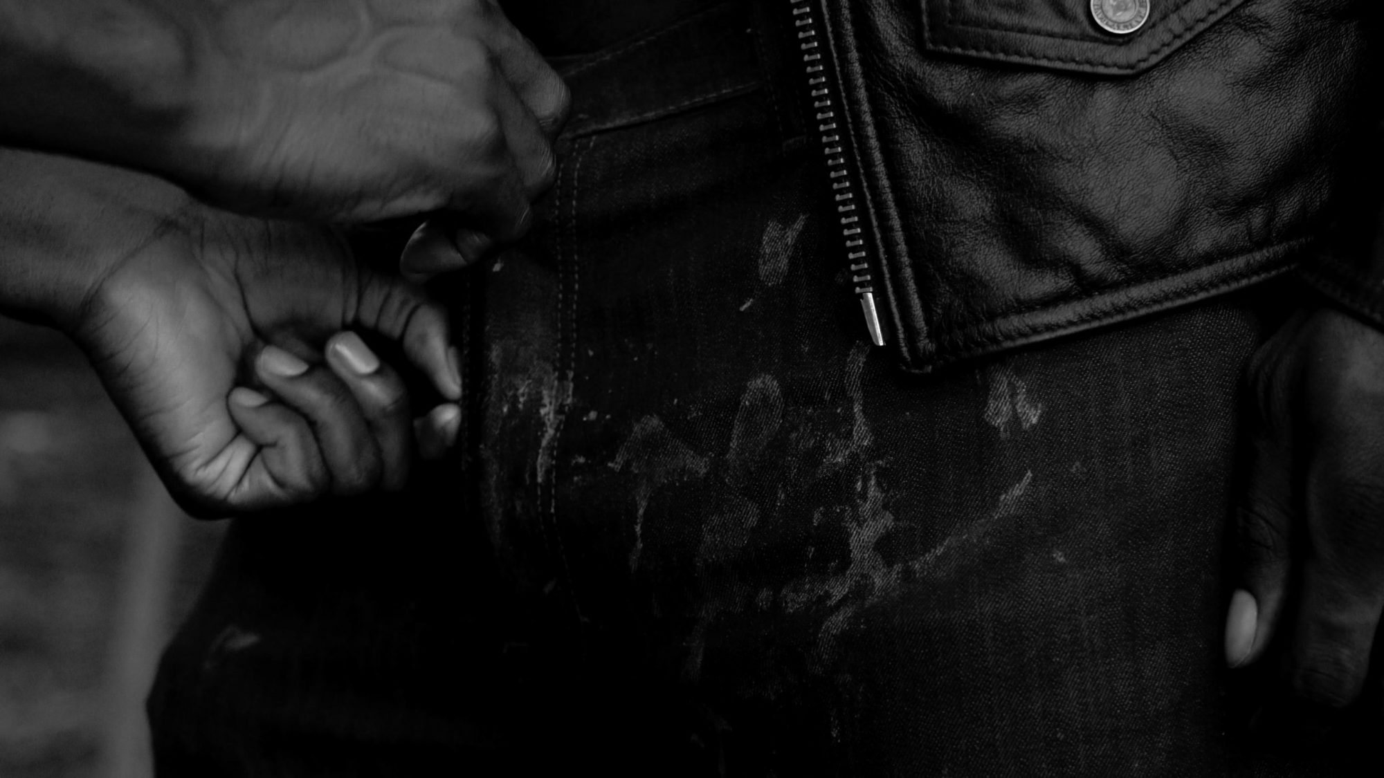 A black and white still of the short film Much Handled Things Are Always Soft. A close up of someone beginning to unzip a man's dirty jeans. He is also wearing a leather jacket.