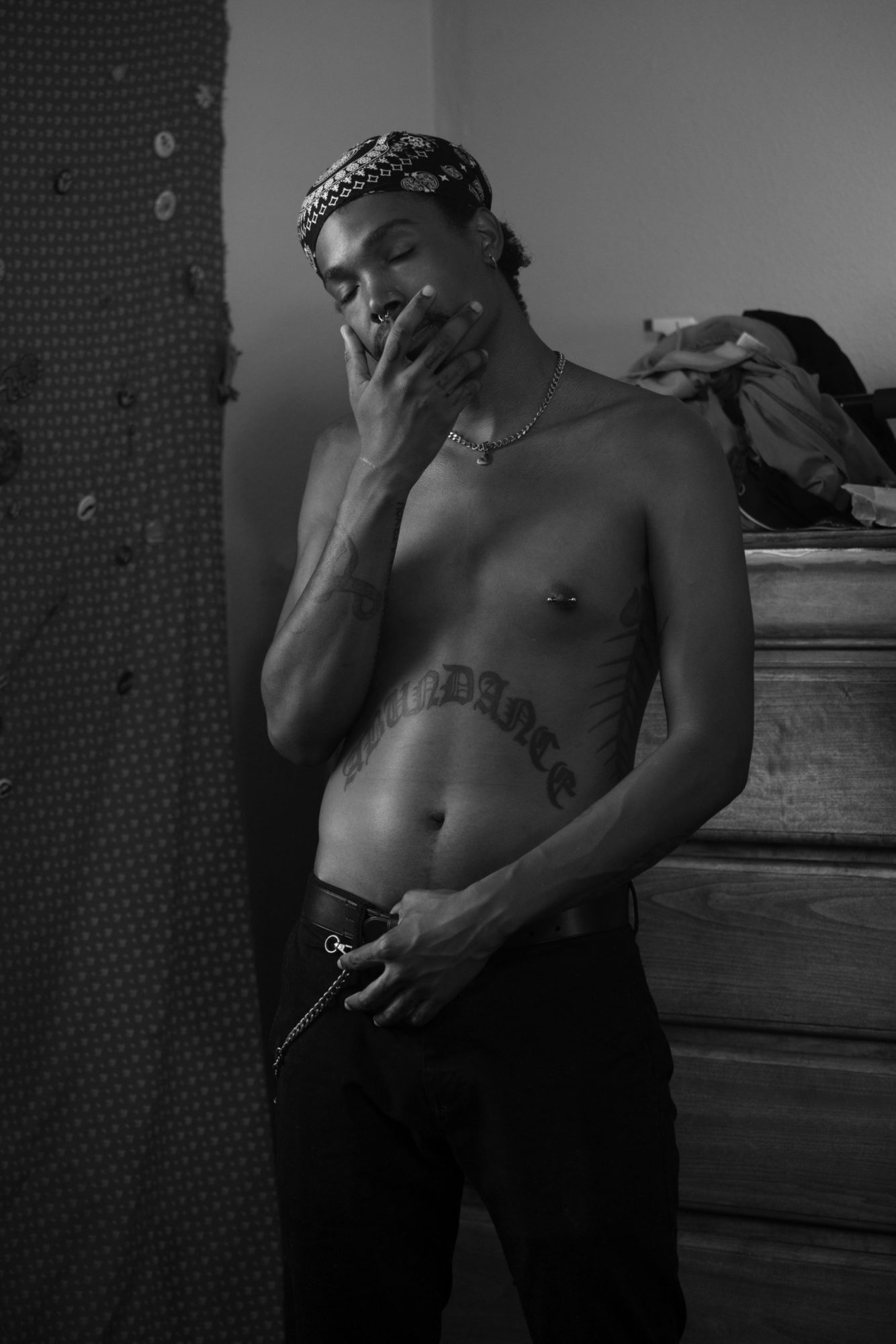 A black and white image of a shirtless man in front of a dresser. He has a tattoo across his abdomen and is wearing belted black jeans. One hand is on the top of his jeans, the other over his mouth. His eyes are closed.