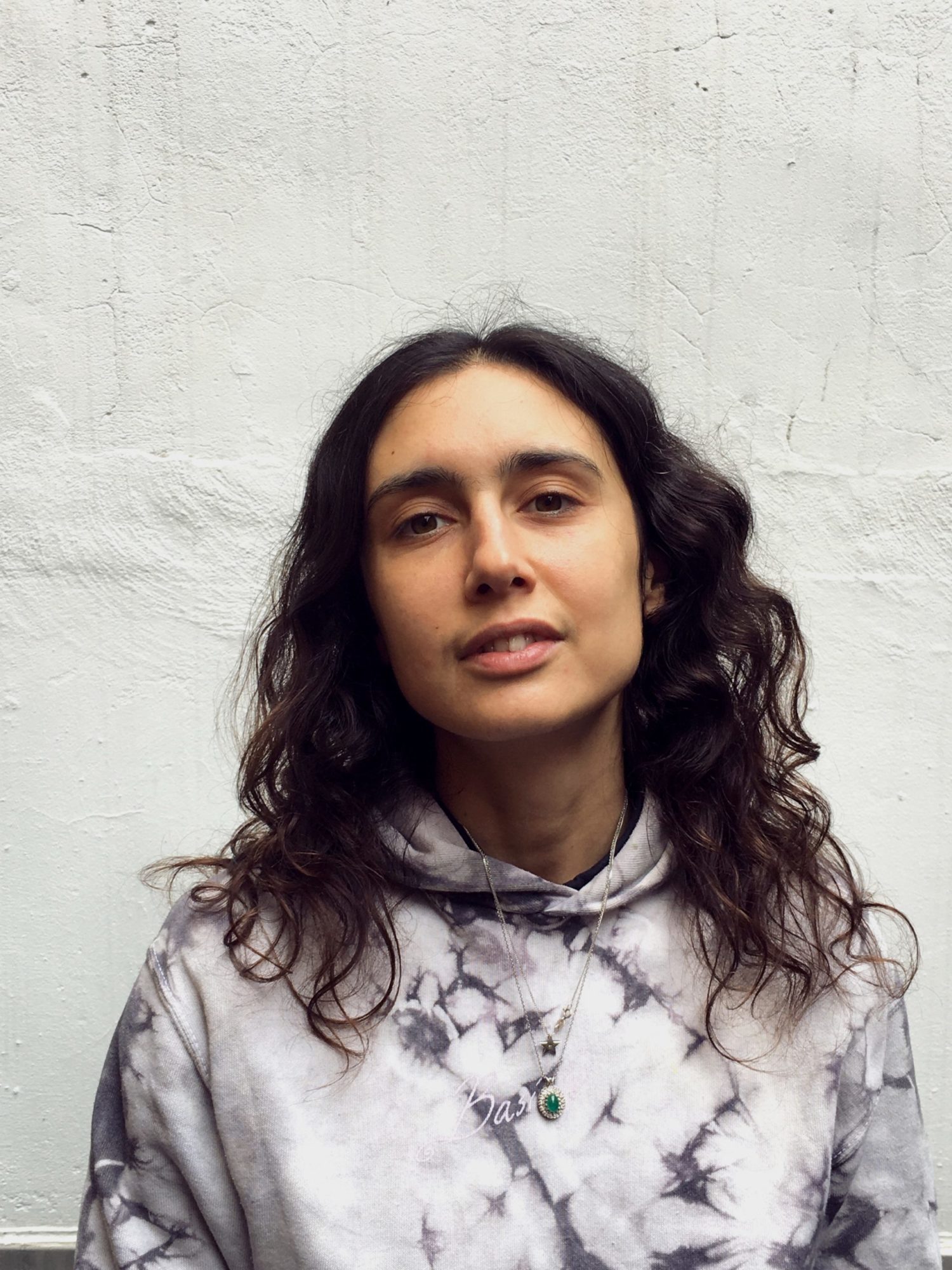 A headshot of a person in a grey and white tie dyed sweatshirt in front of a white plaster wall.