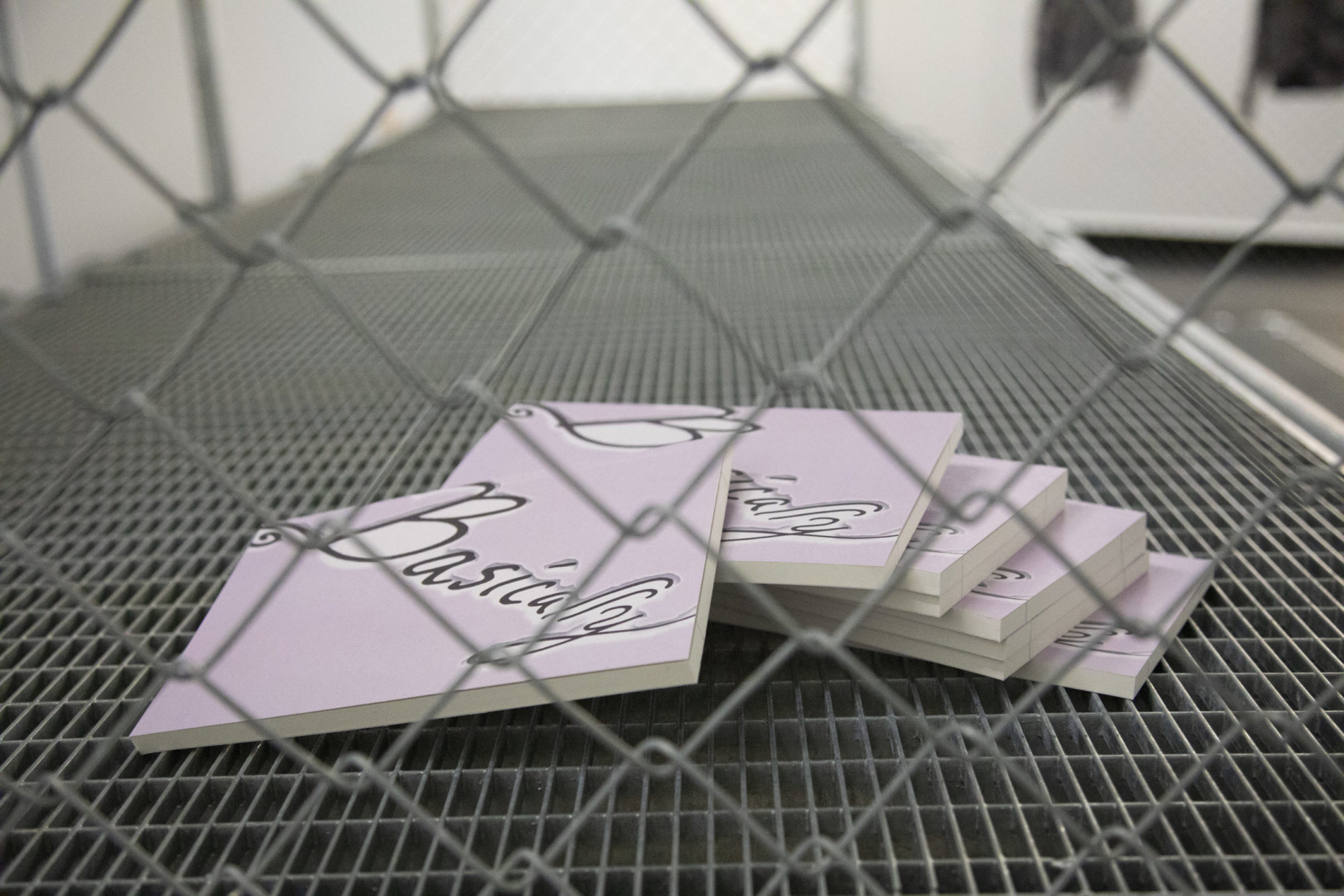 Through a chain link fence, a splayed out stack of lavender squares with basically written on them are in view. There are eight in the stack.