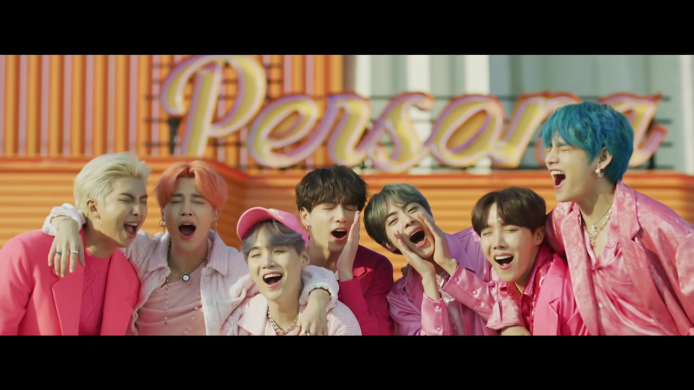 All the members of BTS are clumped together, their arms around one another, with a sign that says persona in the background. They are all singing and wearing pink with happy looks on their faces.