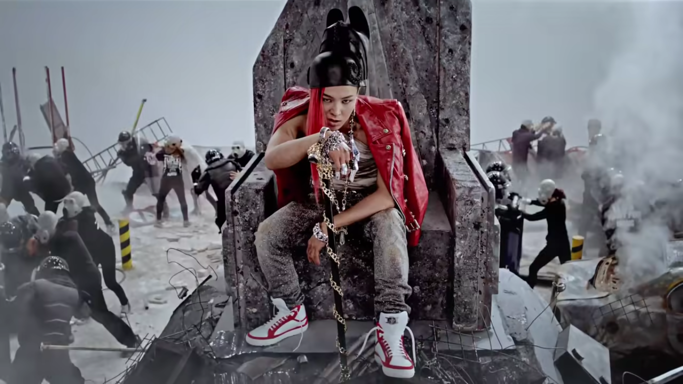 A member of Big Bang sits elevated on a strone throne as a war rages on behind him. He is wearing lots of jewelry and holding a cane. His jacket is read leather and his hair is extremely long and red.