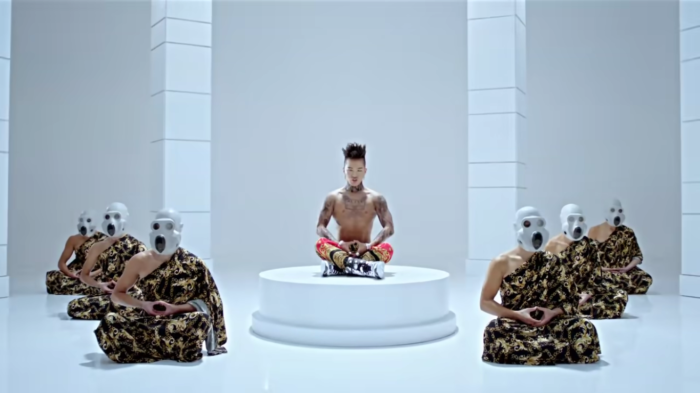 A member of BigBang sits in a white pedestal in an all white room, shirtless, and in position to meditate. Below the pedestal, there are six people wearing patterned robes and white masks resembling gas masks. They are also in position to meditate.