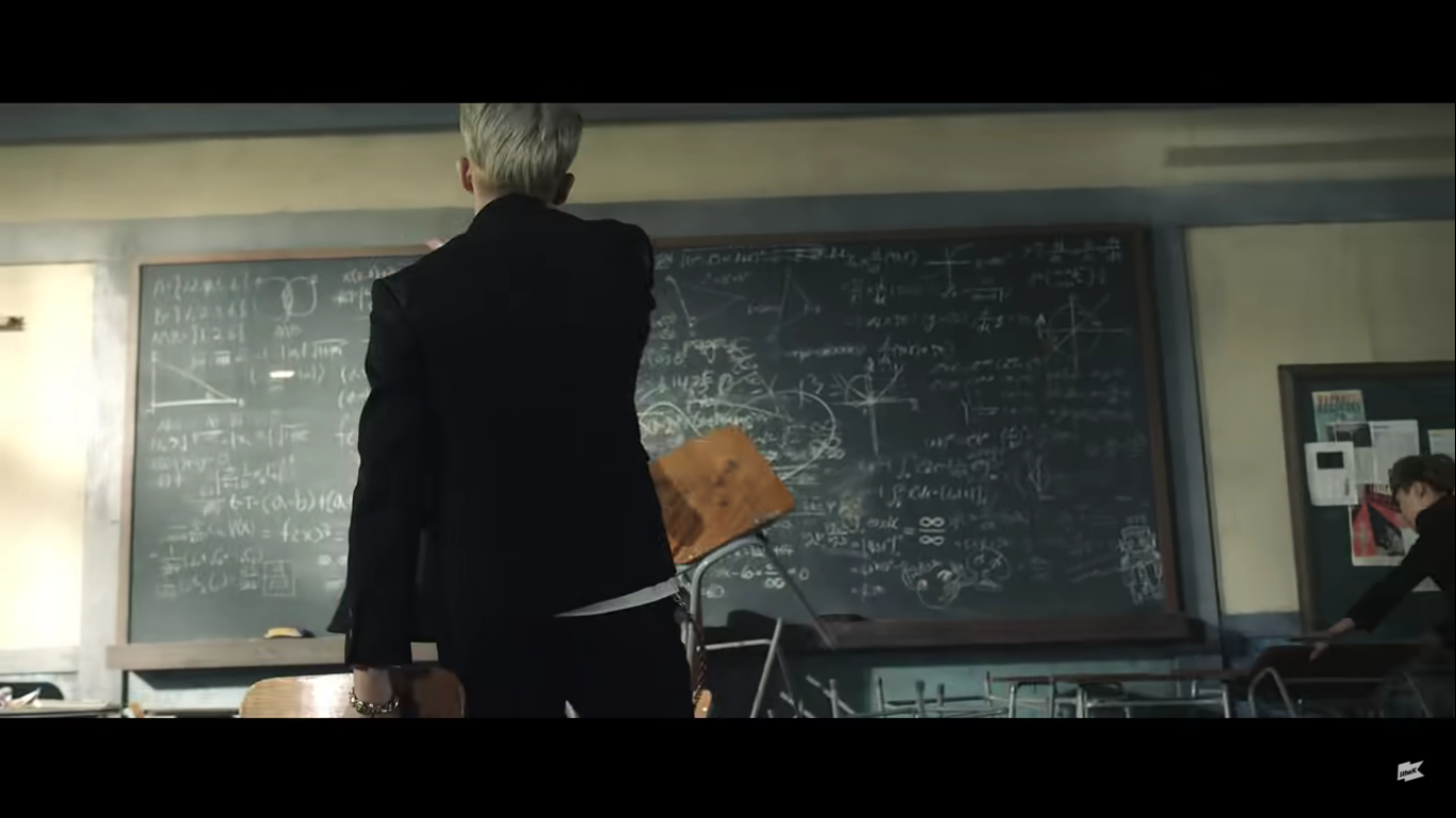 A view from behind of a member of BTS throwing a chair at a chalkboard. The chalkboard is full of math and the chairs and desks are all pushed in front of it.