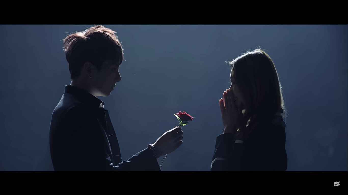 A member of BTS hands a rose to a school girl, her hands are in front of her mouth. They are backlit and the background is solid gray.