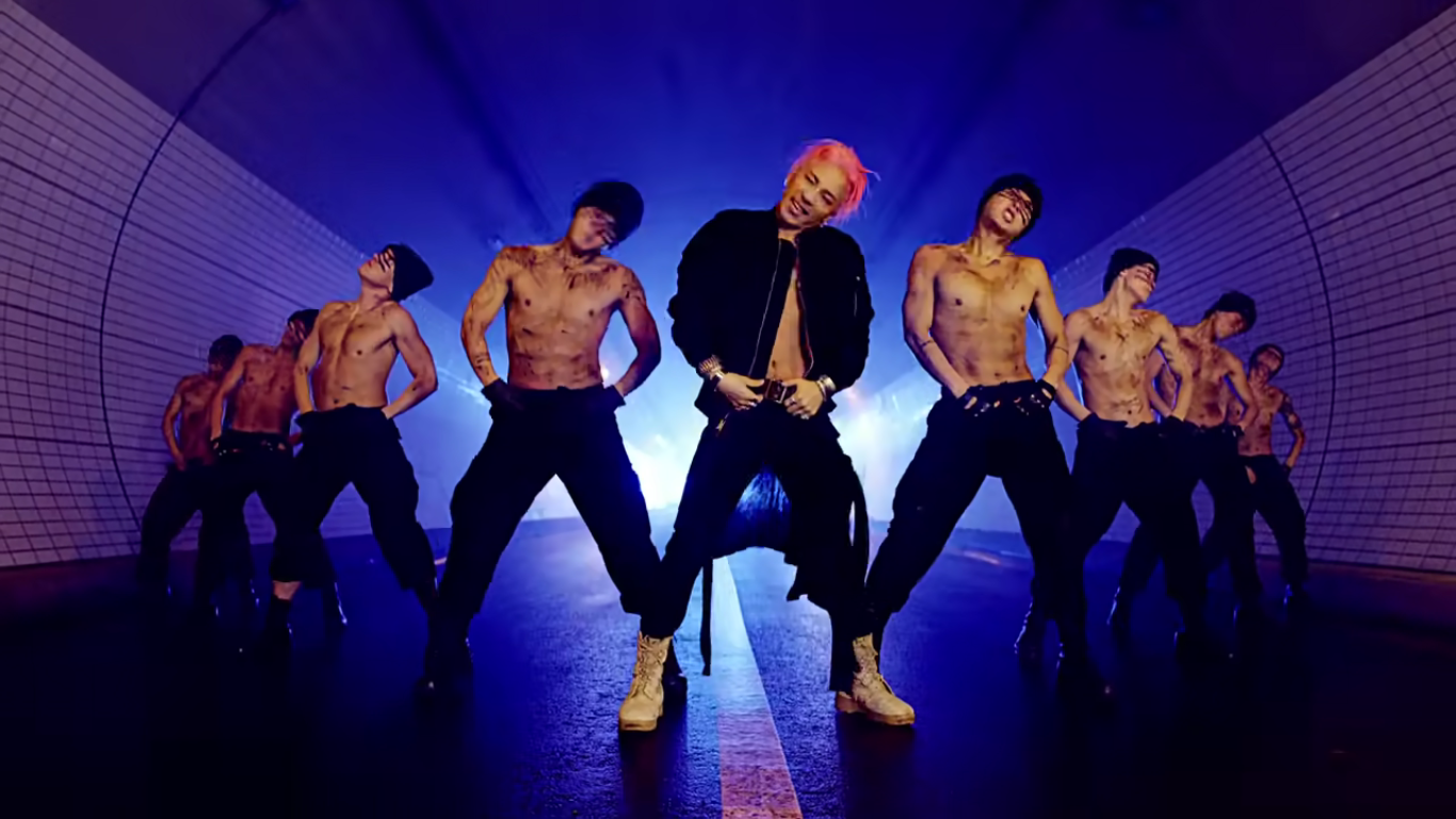 A member of Big Bang leads a group of men dancing in a road tunnel. They are all positioned in a V position, the member of Big Bang at the front.. All the men are shirtless and wearing black pants. The member of Big Bang is also wearing a jacket and has pink hair. All the other men are wearing black beanies and have black stripes across their eyes.