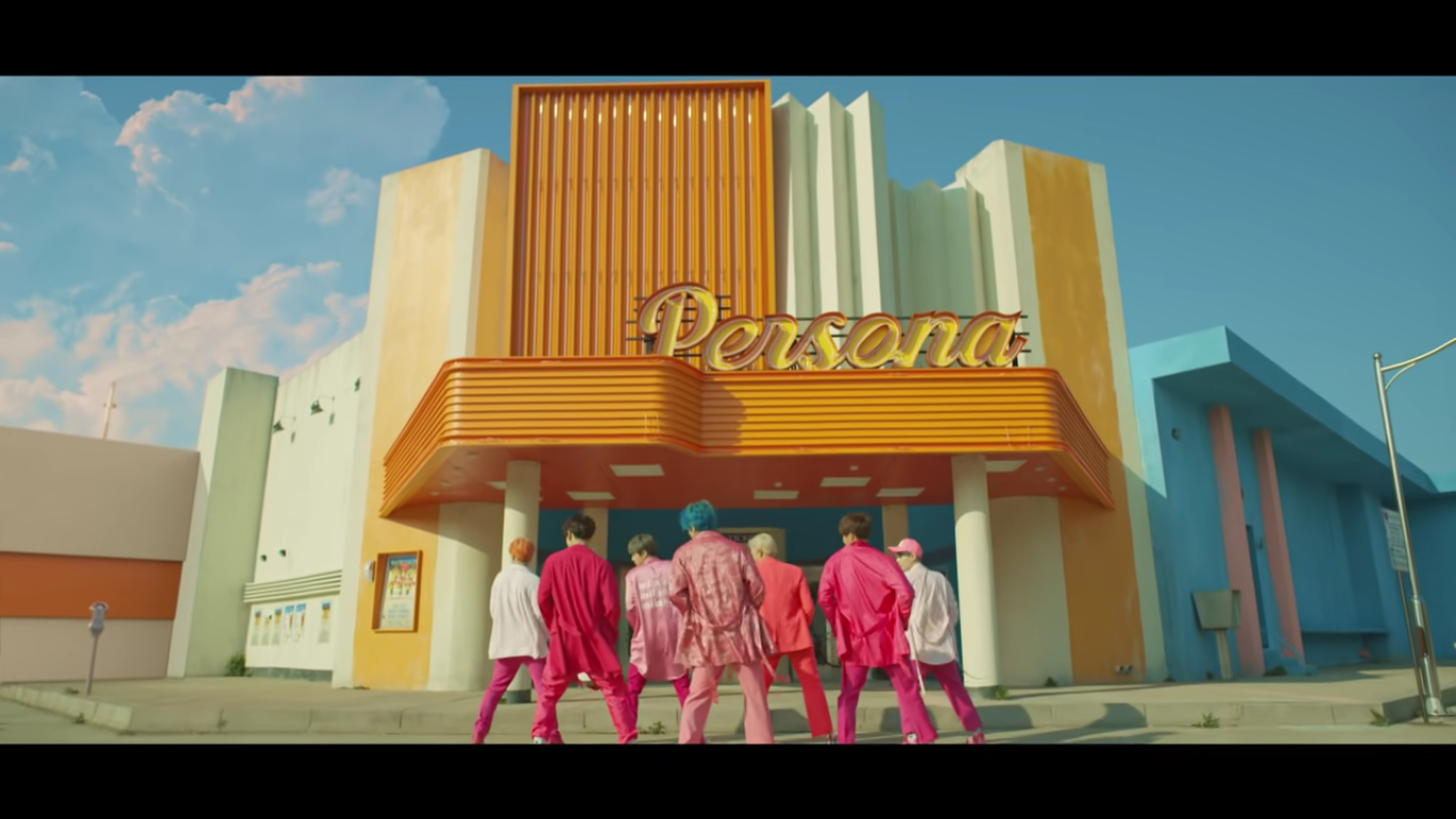 The members of BTS stand staggered so all are in view, all their backs facing the camera and caught in the same position mid dance. They are all wearing different shades of pink in front of a retro looking theatre named 'Persona'. The theatre is white, yellow, and blue.