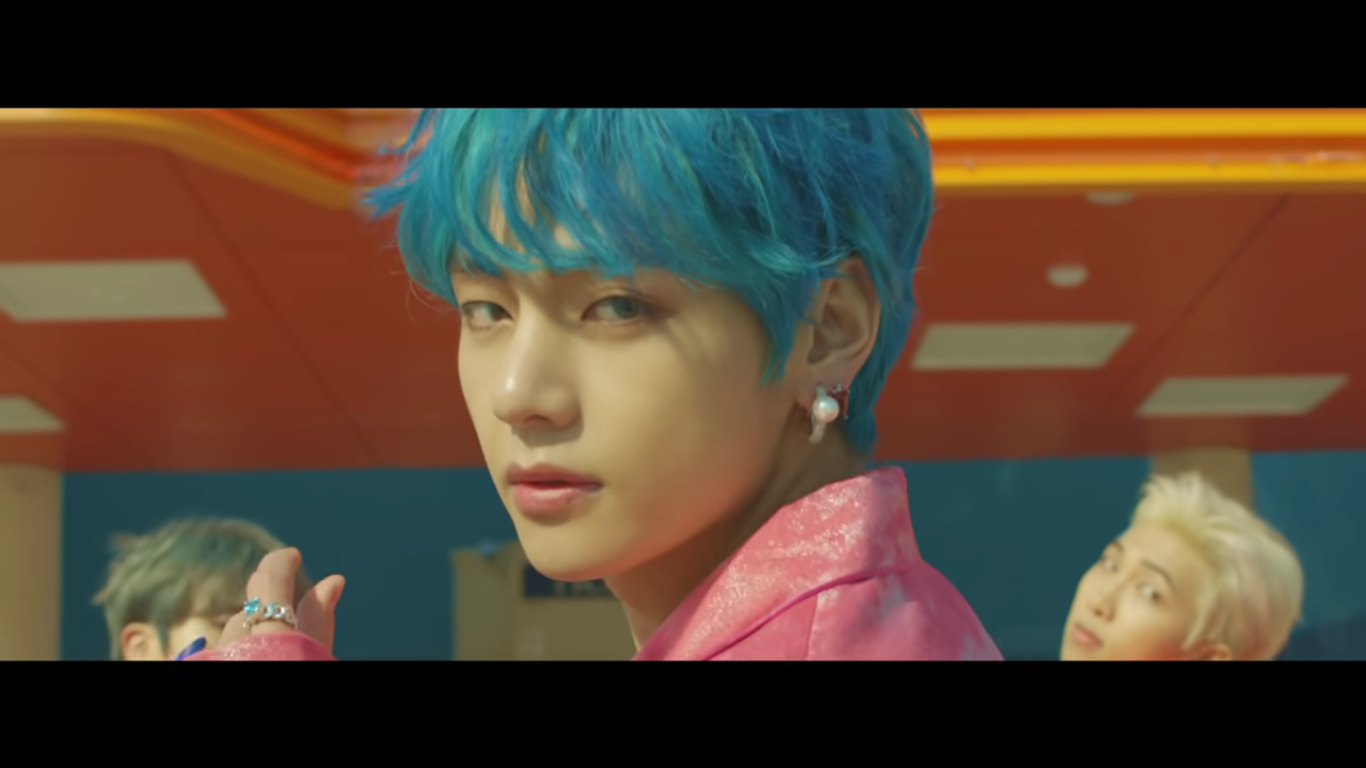 A closeup of one of the members of BTS' face. He ahs blue hair and has turned to face the camera. Two other members are in sight in the background.