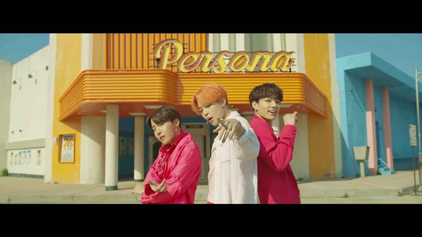 Three members of BTS stand facing the camera, the one in the center pointing at the camera. They are all wearing different shades of pink in front of a retro looking theatre named 'Persona'. The theatre is white, yellow, and blue.