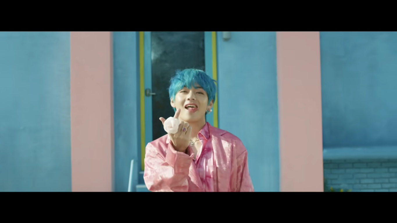 A solo shot of a member of BTS. He is facing the camera, smiling, and pointing up. His hair is blue and his outfit is light pink; the wall he is in front of is also blue and light pink.