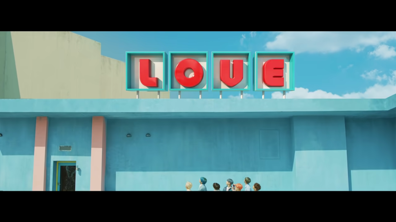 The members of BTS, barely in view at the bottom of the shot, look up to a sign that says LOVE in retro ltters. The building holding the sign is blue, the letters are yellow in blue and white boxes.