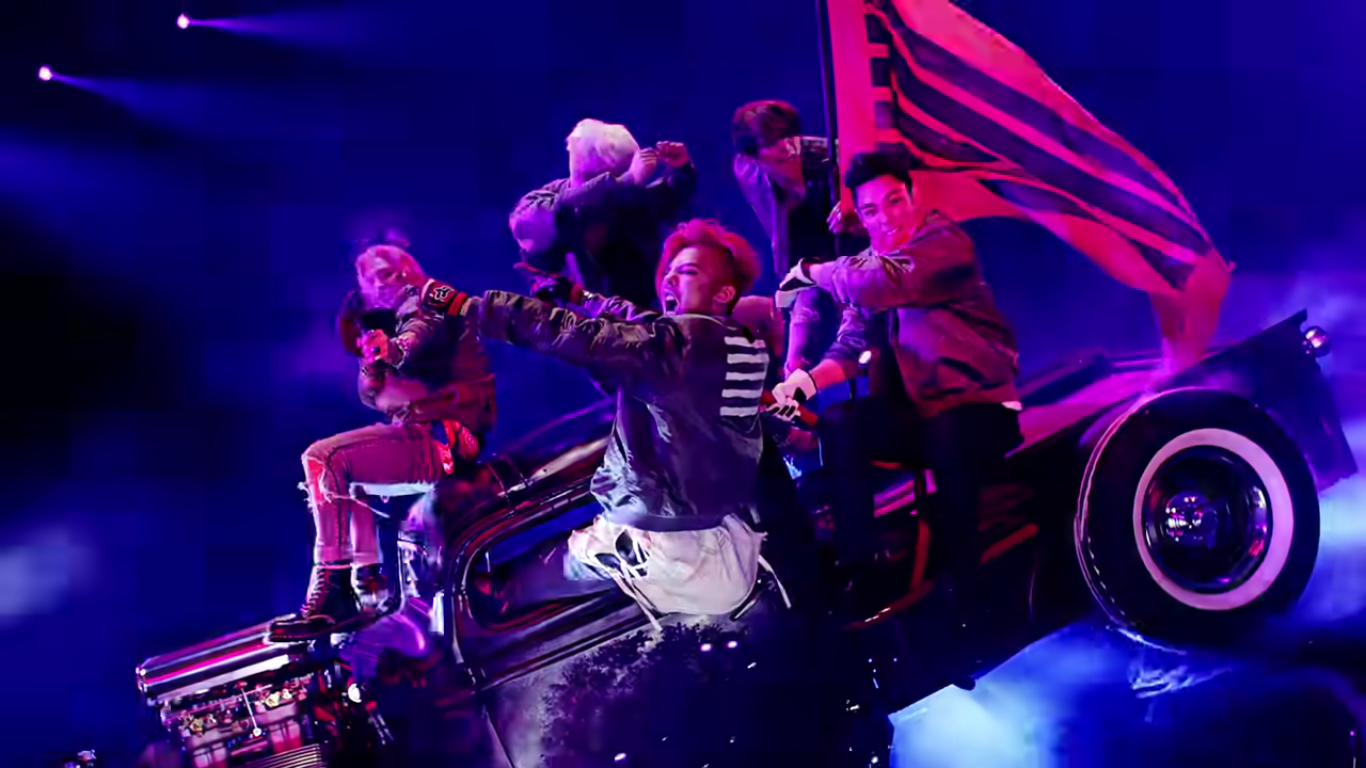 The members of Big Bang are seated on a truck together, all of them dancing and yelling. They are wielding a red flag with black stripes.