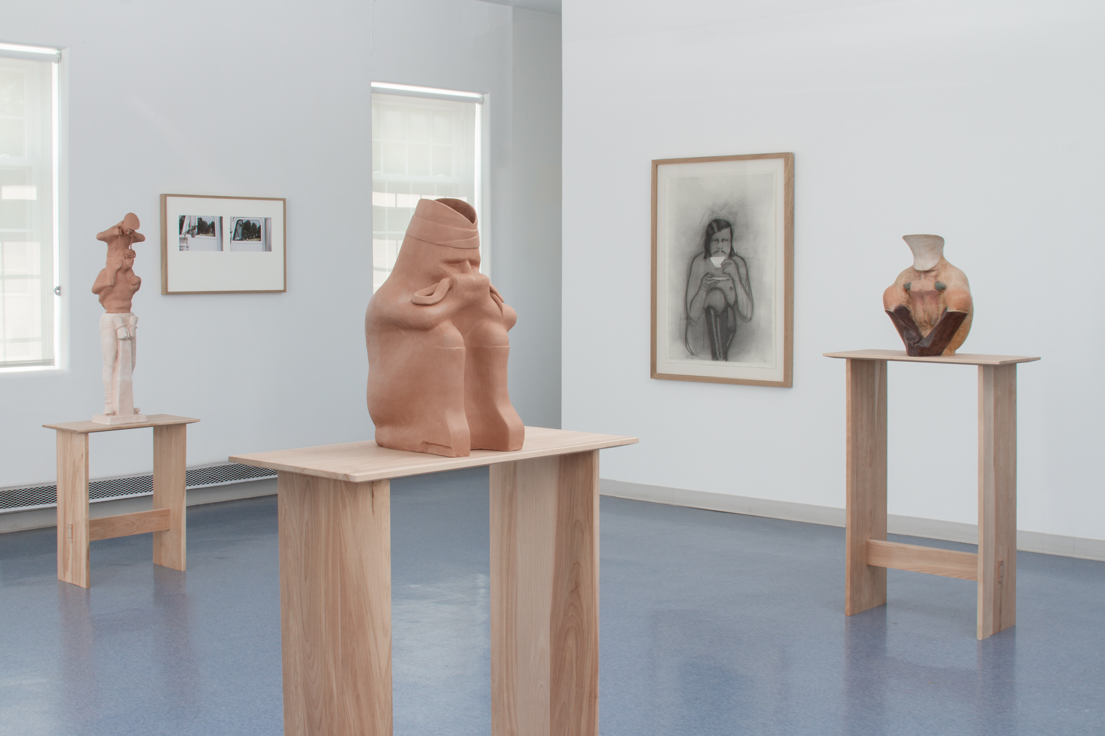 A gallery view of the show. There are three sculptures on wooden tables. There are two photographs in one wooden frame and a charcoal drawing in another.