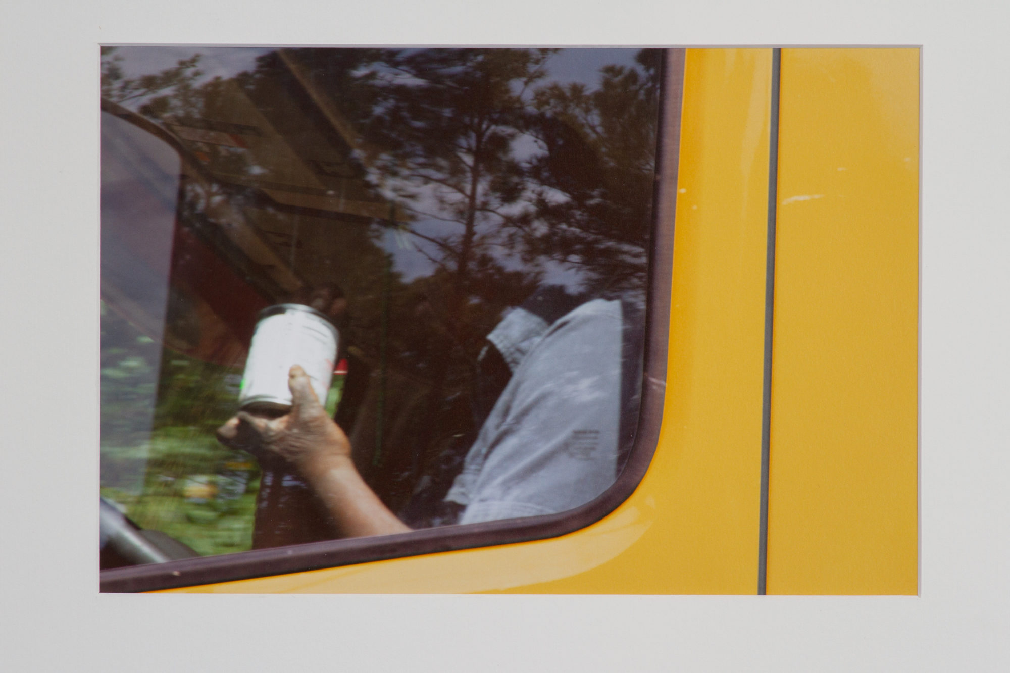 An angled up view of a truck window. The truck is yellow. A person's arm is in view but their face is in the shadows. They are holding a can. The view is distorted by the reflection of trees.