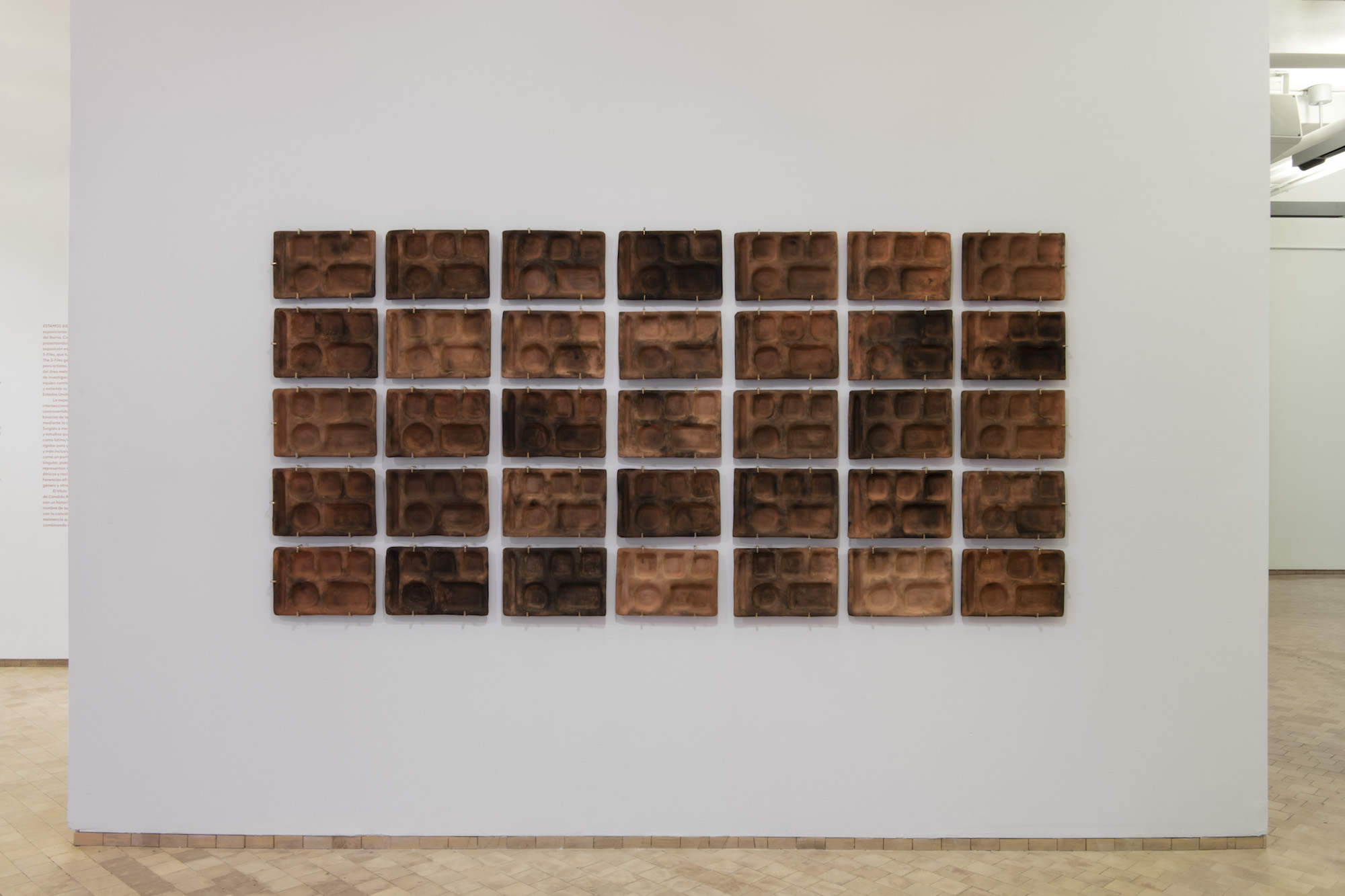 Art Installation is presented on white gallery walls. Various brown ceramic trays are displayed in a rectangular grid.
