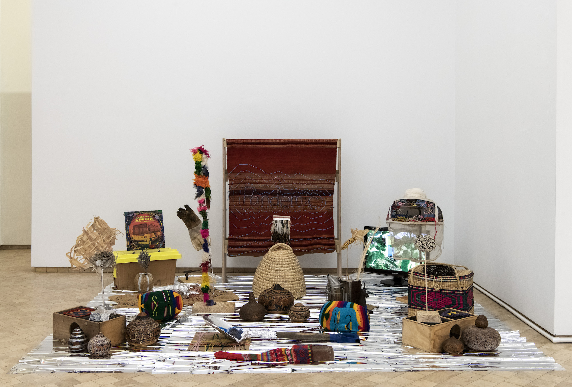 Found objects are scattered around a survival blanket. Bee keeping equipment, a tapestry, and dried plants are among the variety of found objects.