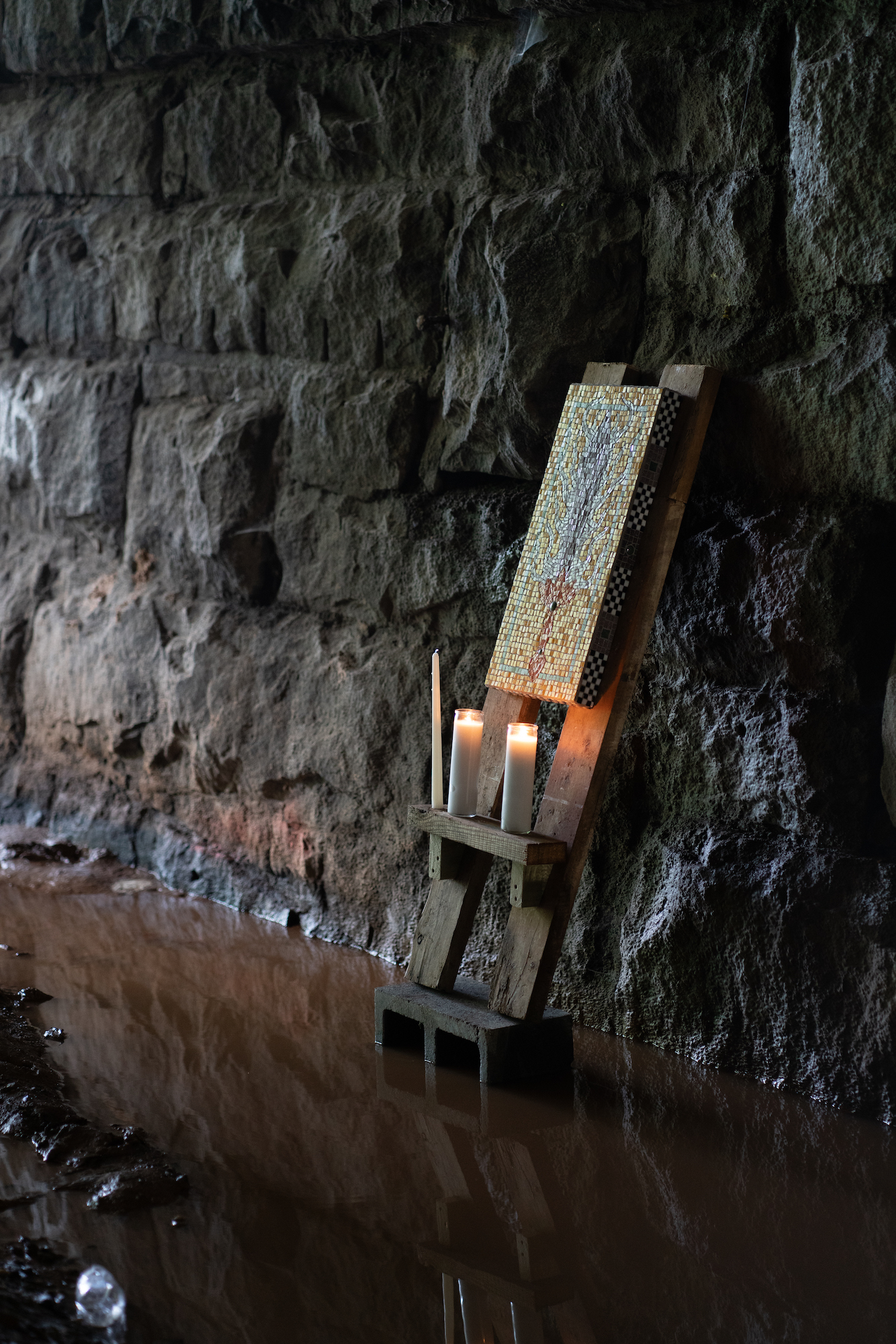 A canvas is propped up on a wooden frame which leans against the wall of a tunnel. The painting is lit from below by two votive candles. The wooden frame is stands in a muddy puddle.