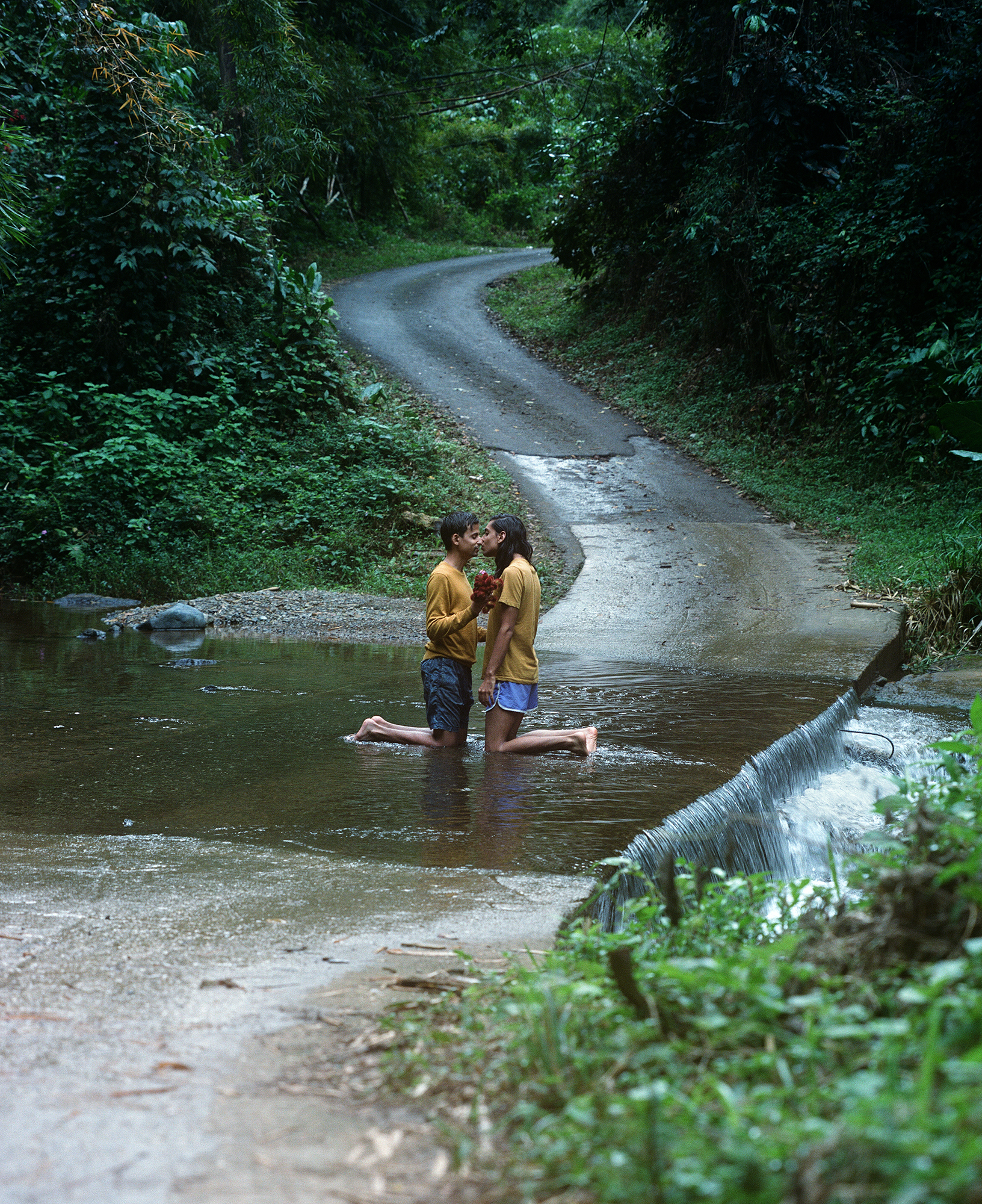 A photo of two male presenting lovers kneeling in small stream is shown. The two are facing each other, as if caught in an intimate moment.