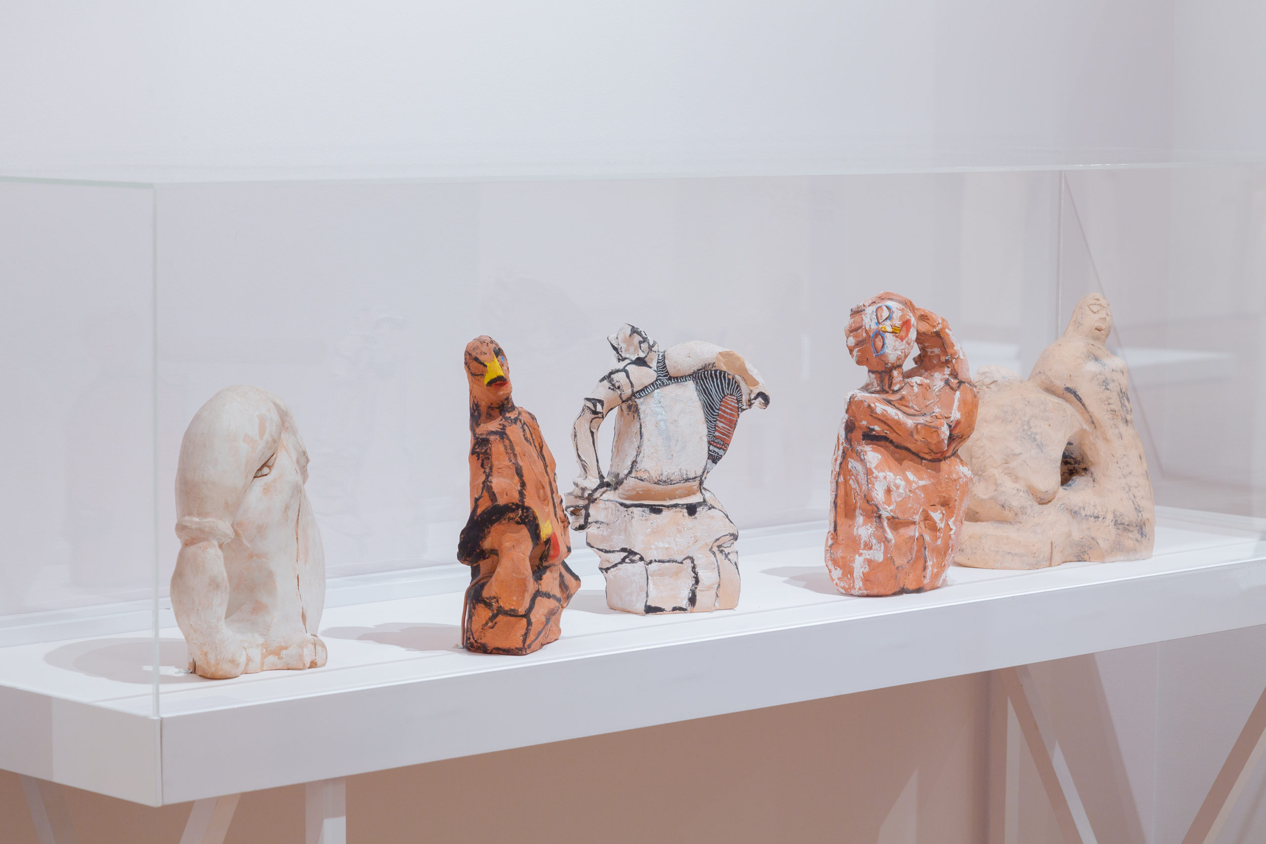 small clay sculptures are presented in a rectangular glass case against white gallery walls.