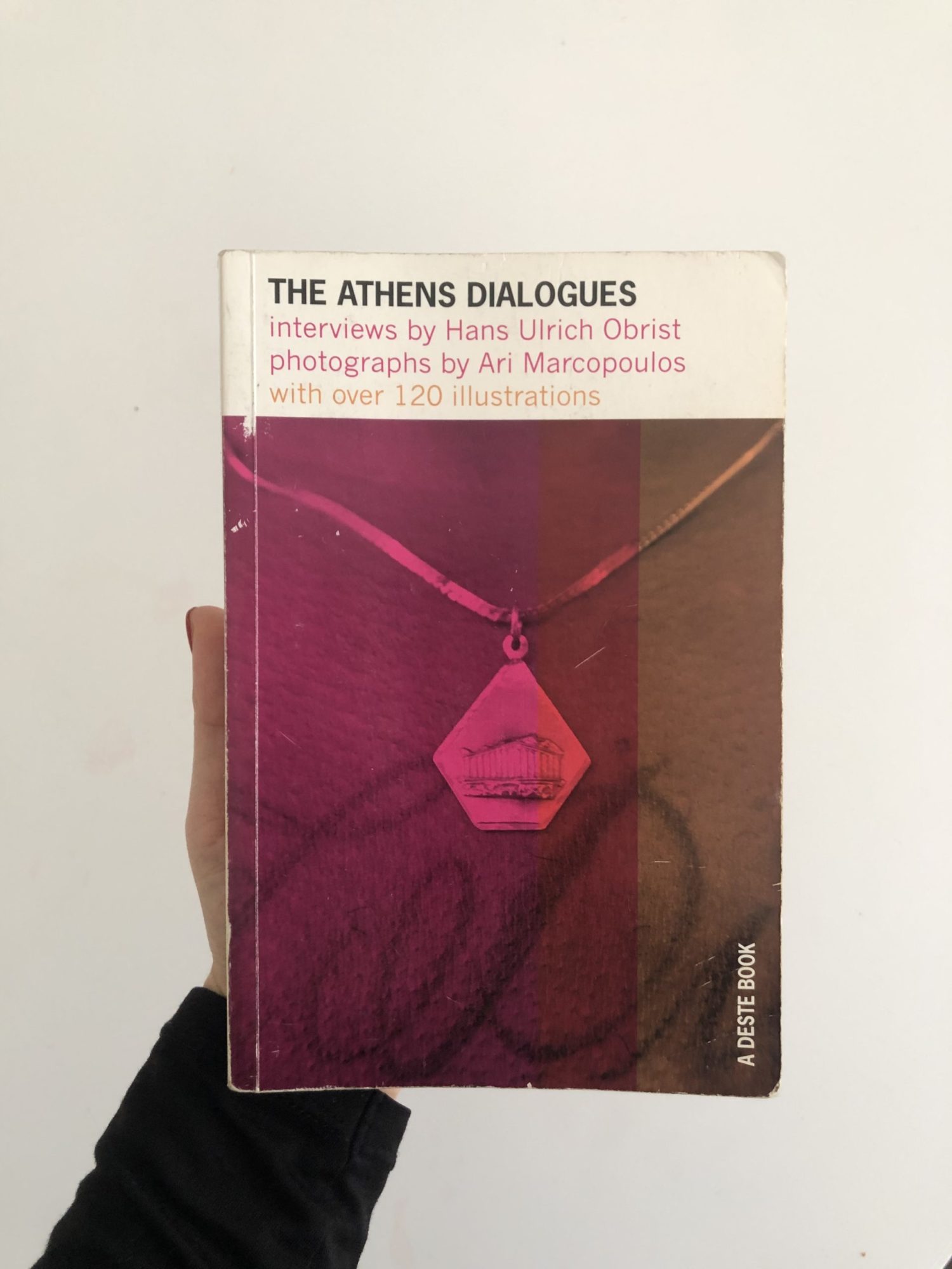 A photo of the front cover of The Athens Dialogues: Interviews by Hans Ulrich Obrist. The cover is pink and orange, and features the title and author at the top.
