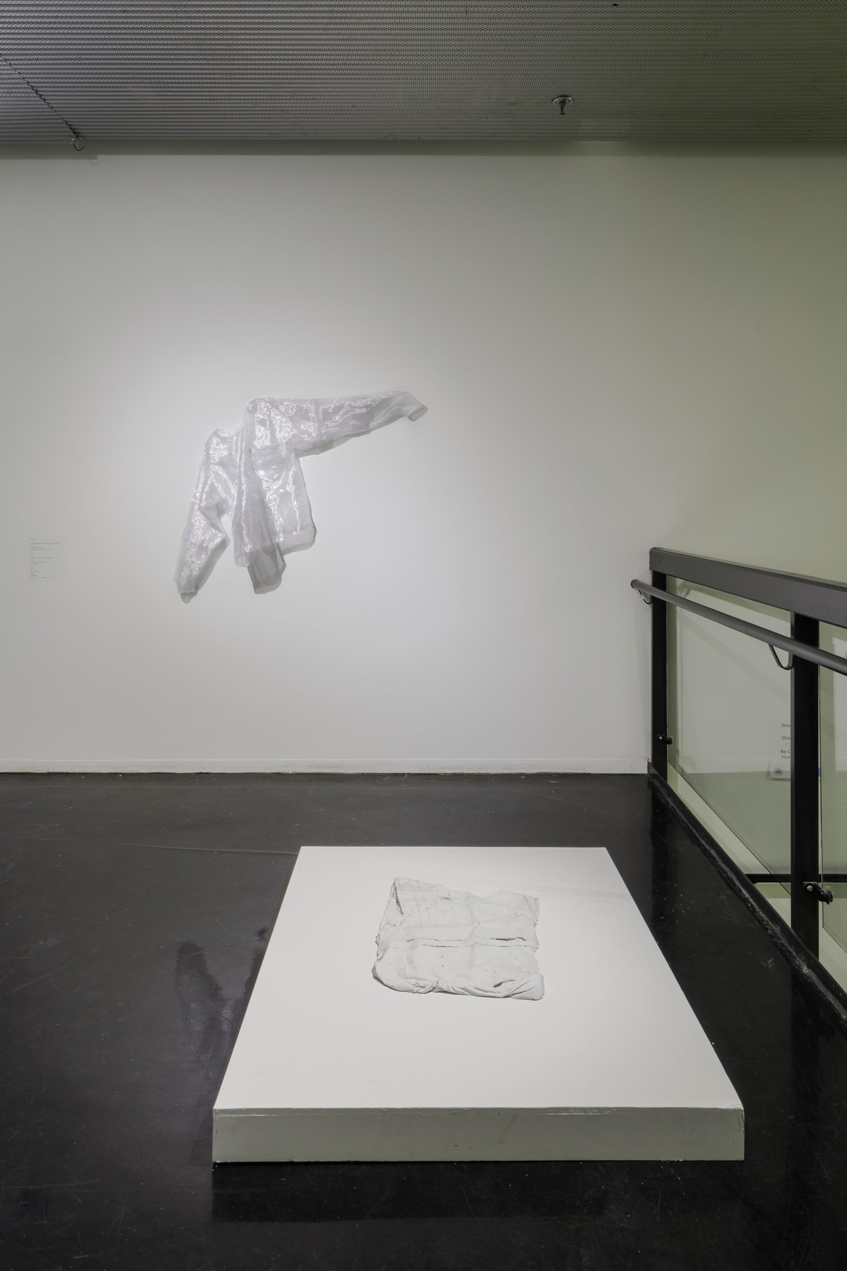 a transparent safety coat is hung on white installation walls while a garment is displayed on the floor on a white installation background.