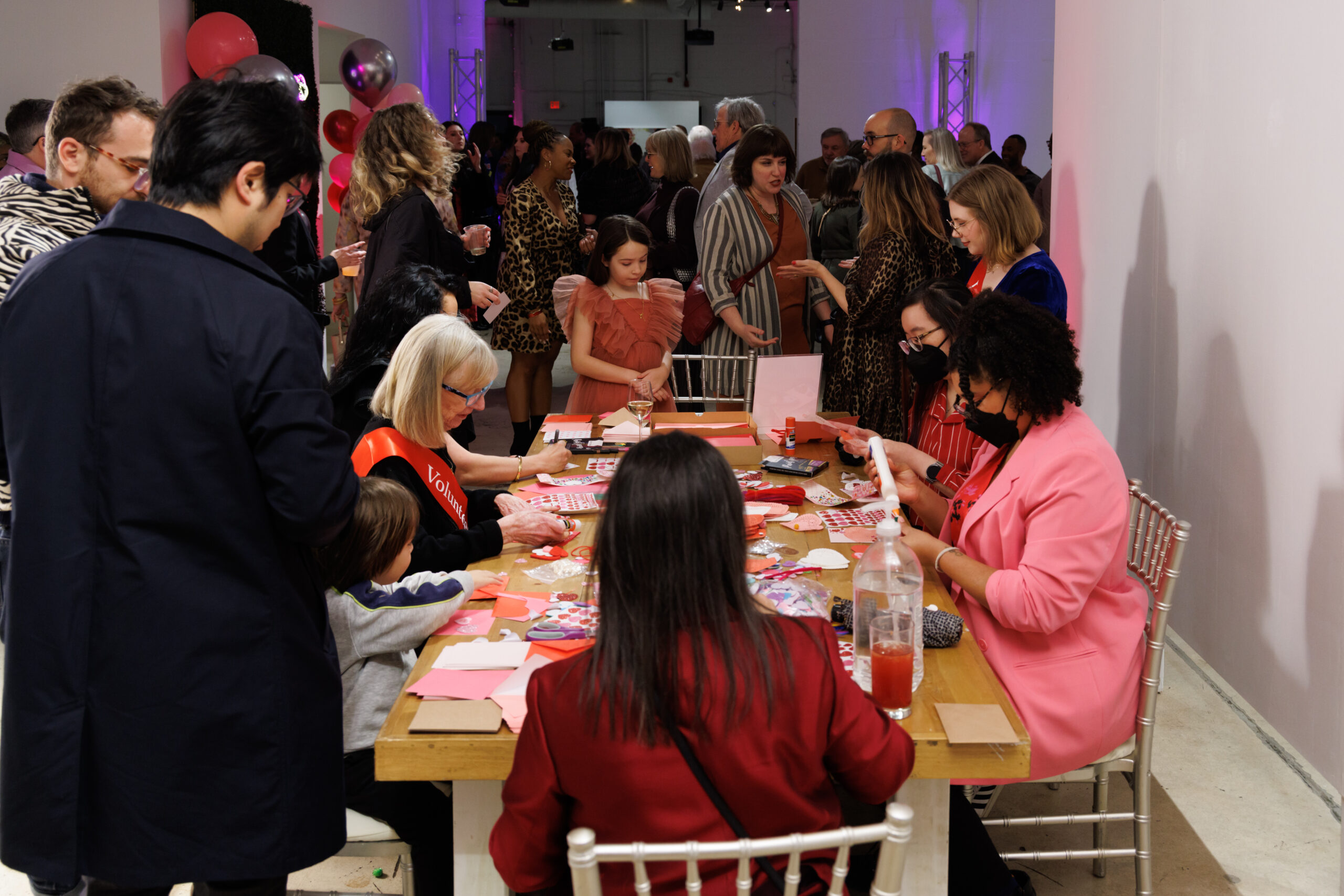 Image of several people sitting at a table with many pieces of pink, red, and white paper in front of them. In the background is a large group of people.
