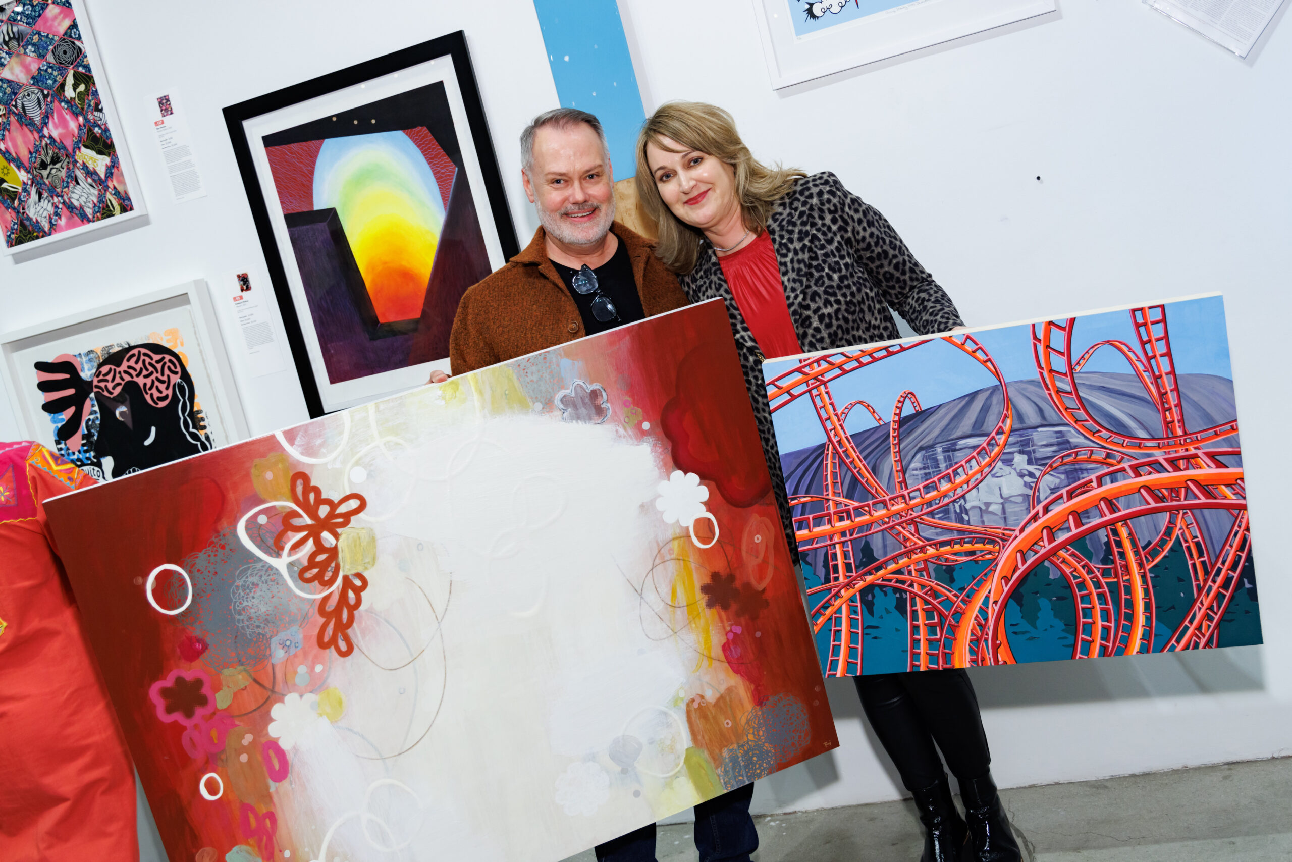 Image of two people posing with two artworks in front of a wall featuring many other artworks. The man is holding a red and white piece with flowers, while the woman is holding a blue and red piece with loops of rollercoaster track.