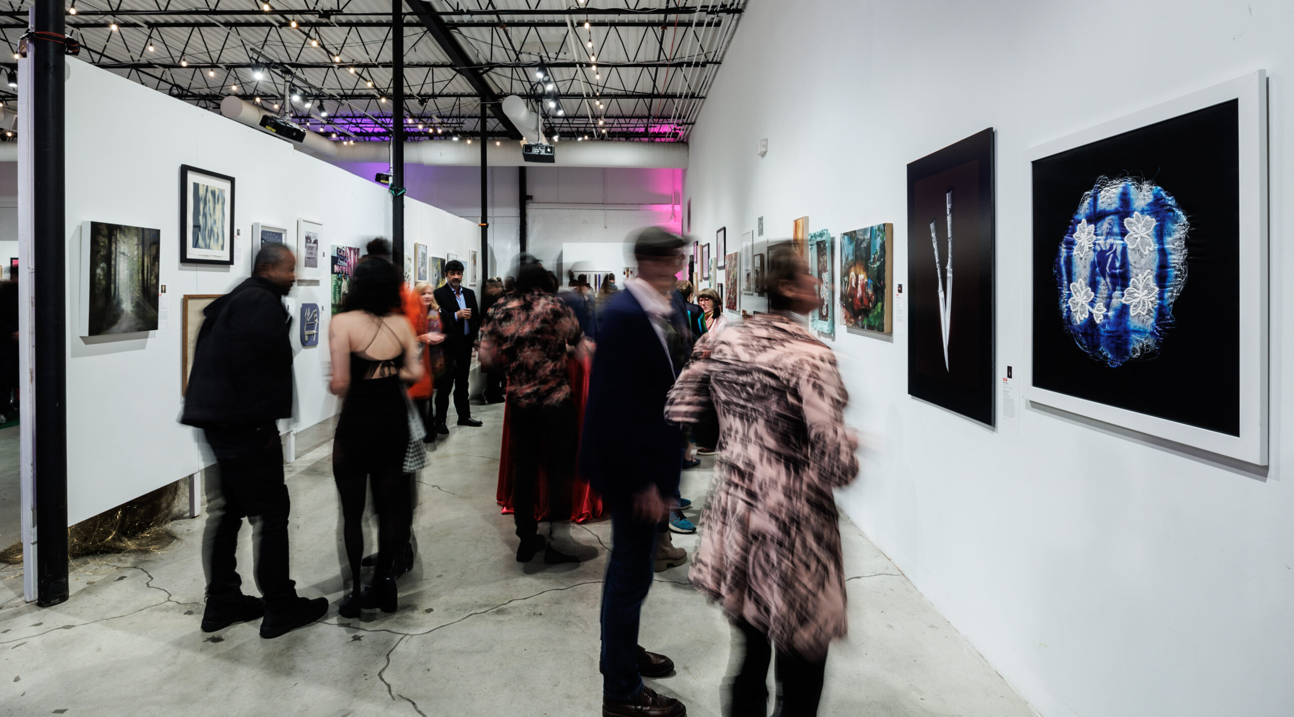 Image of a crowd of people, some blurred, talking to each other and looking at many different framed artworks on white walls.