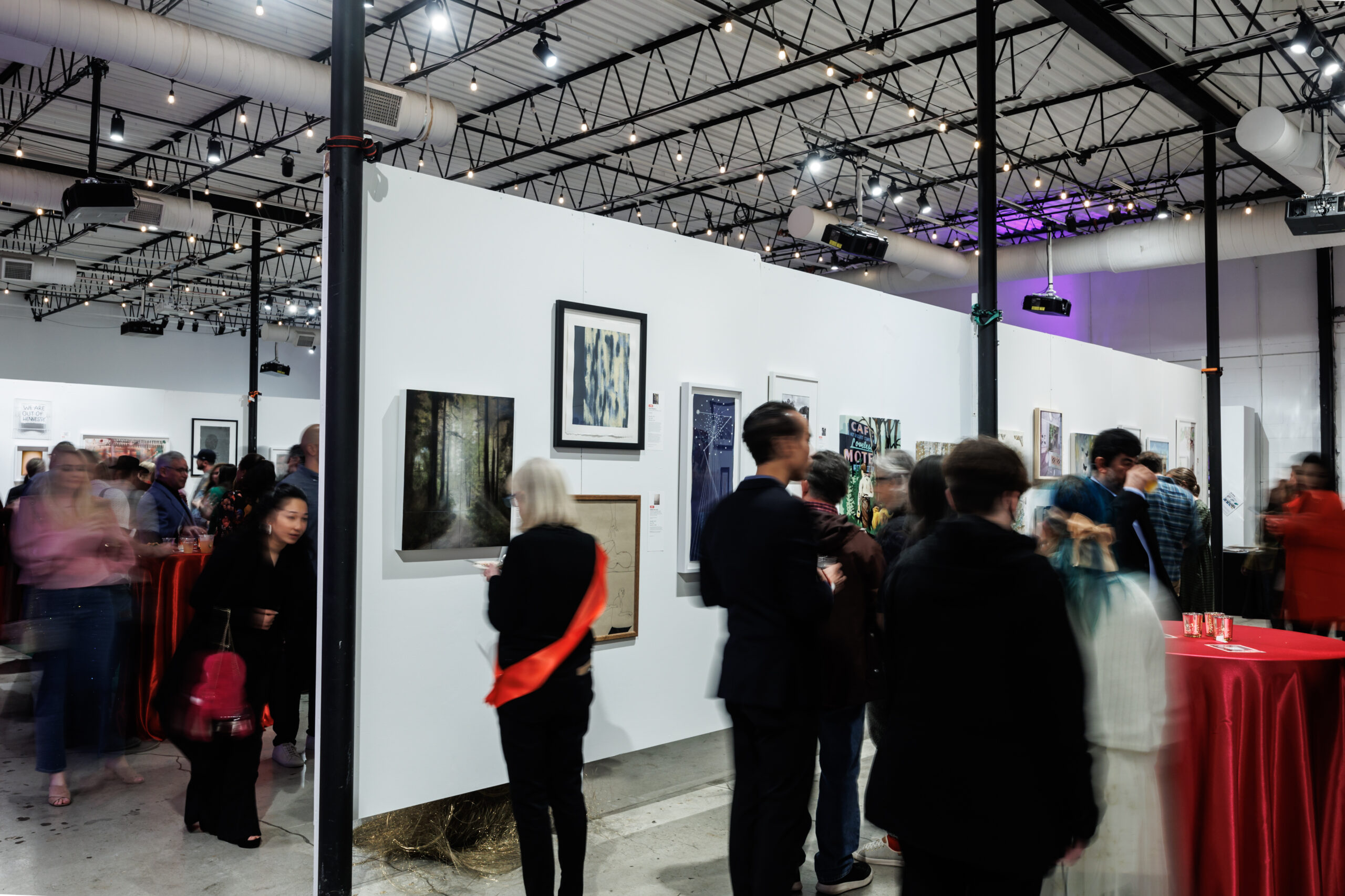 Image of a crowd of people, some blurred, talking to each other and looking at many different framed artworks on white walls.
