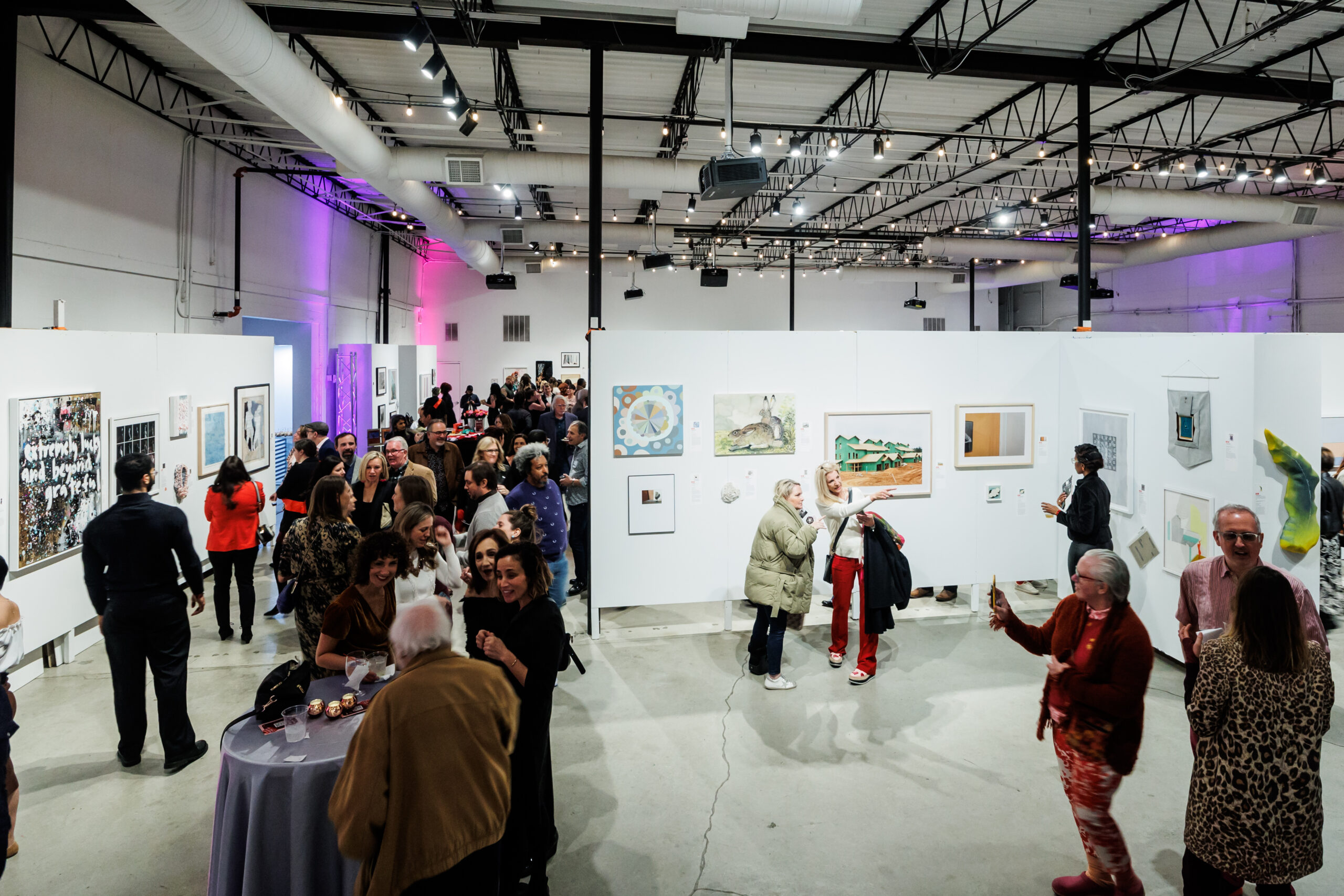 Image of a crowd of people talking to each other and looking at many different framed artworks on white walls.