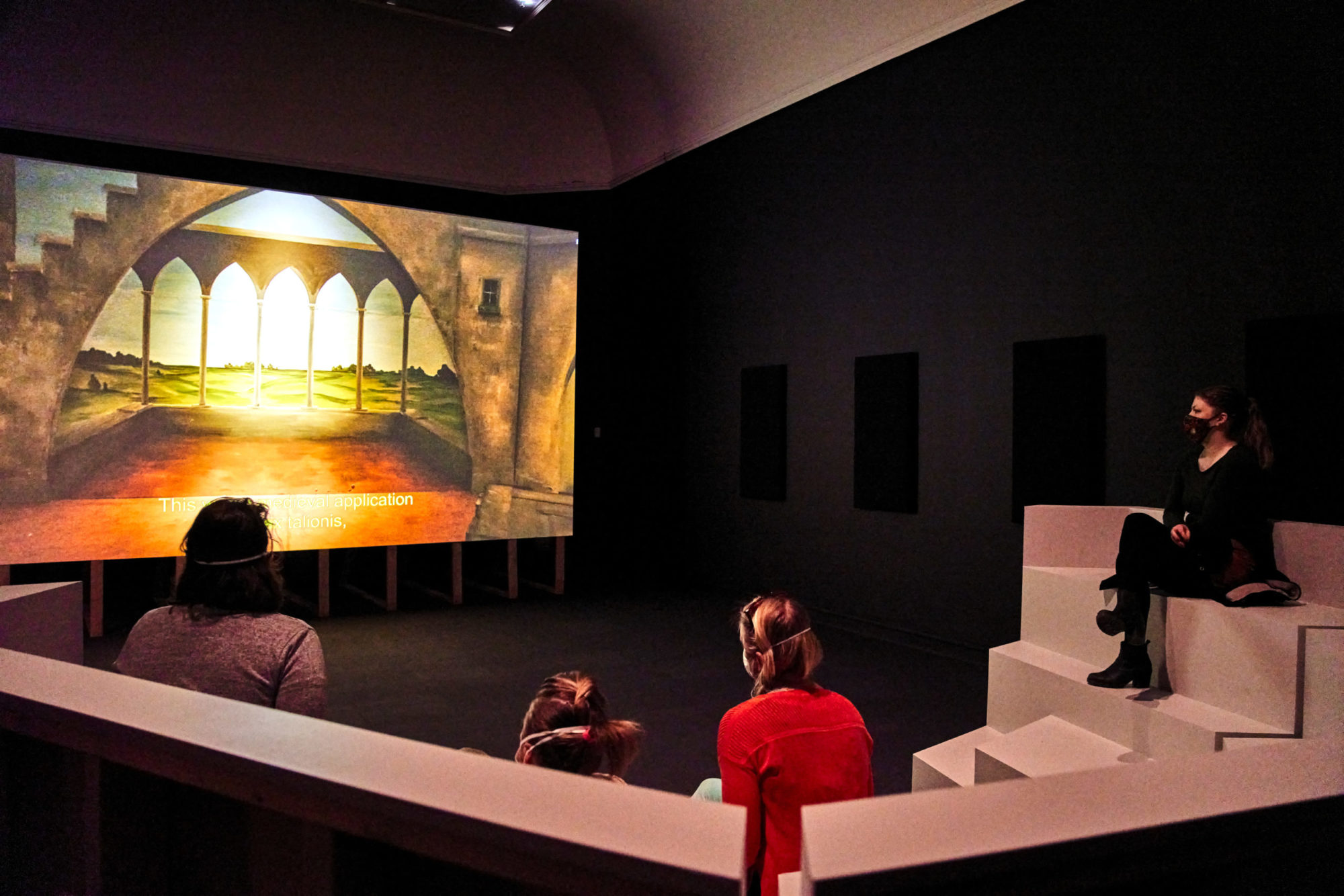 Installation view of Bugs & Beasts Before the Law. Audience members sit on a set of stairs. They watch a screen that is on a still of pointed Gothic arches.